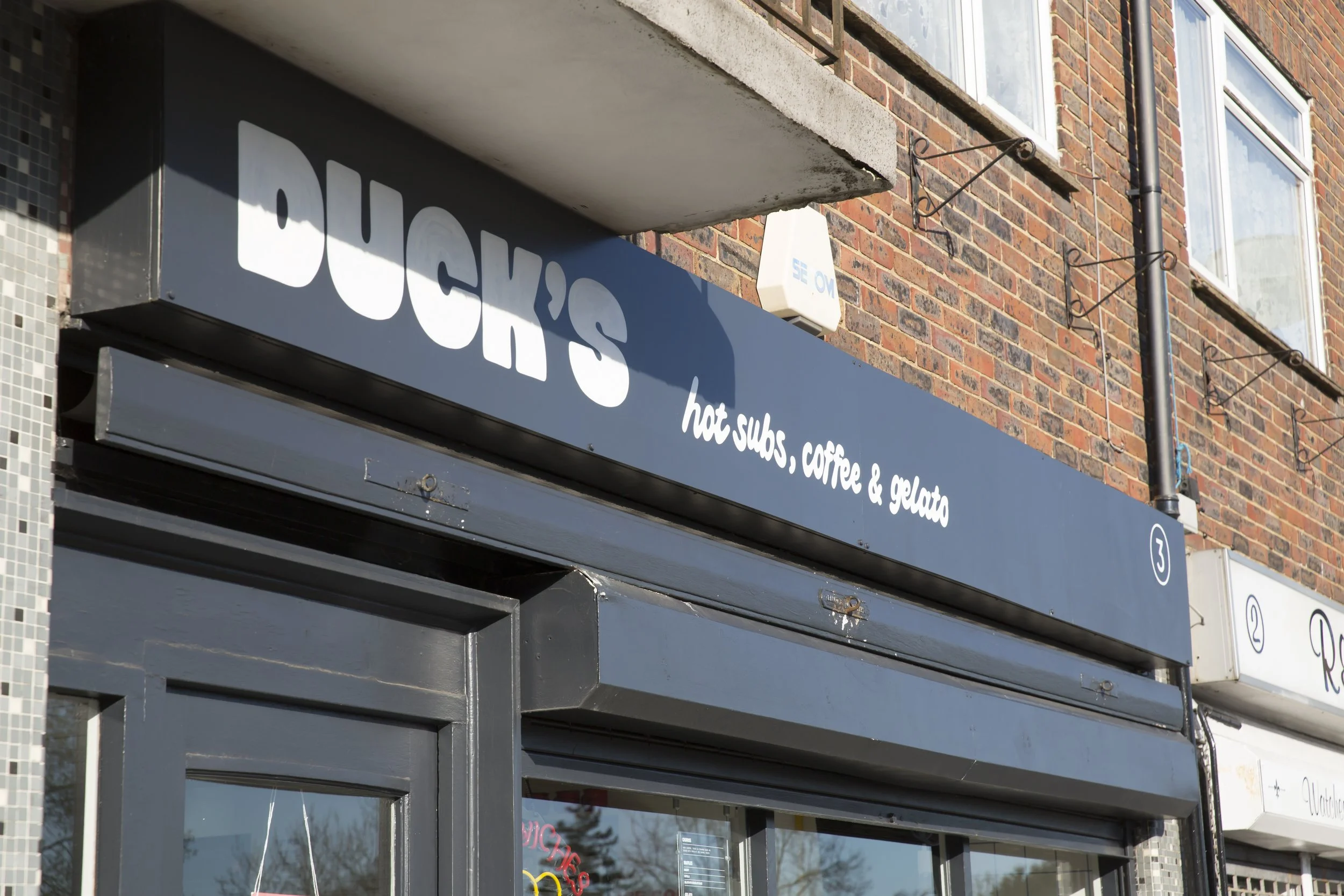 Storefront with a black sign that reads 'DUCKY'S' in large white letters, offering hot subs, coffee, and gelato, with a brick building and windows in the background.