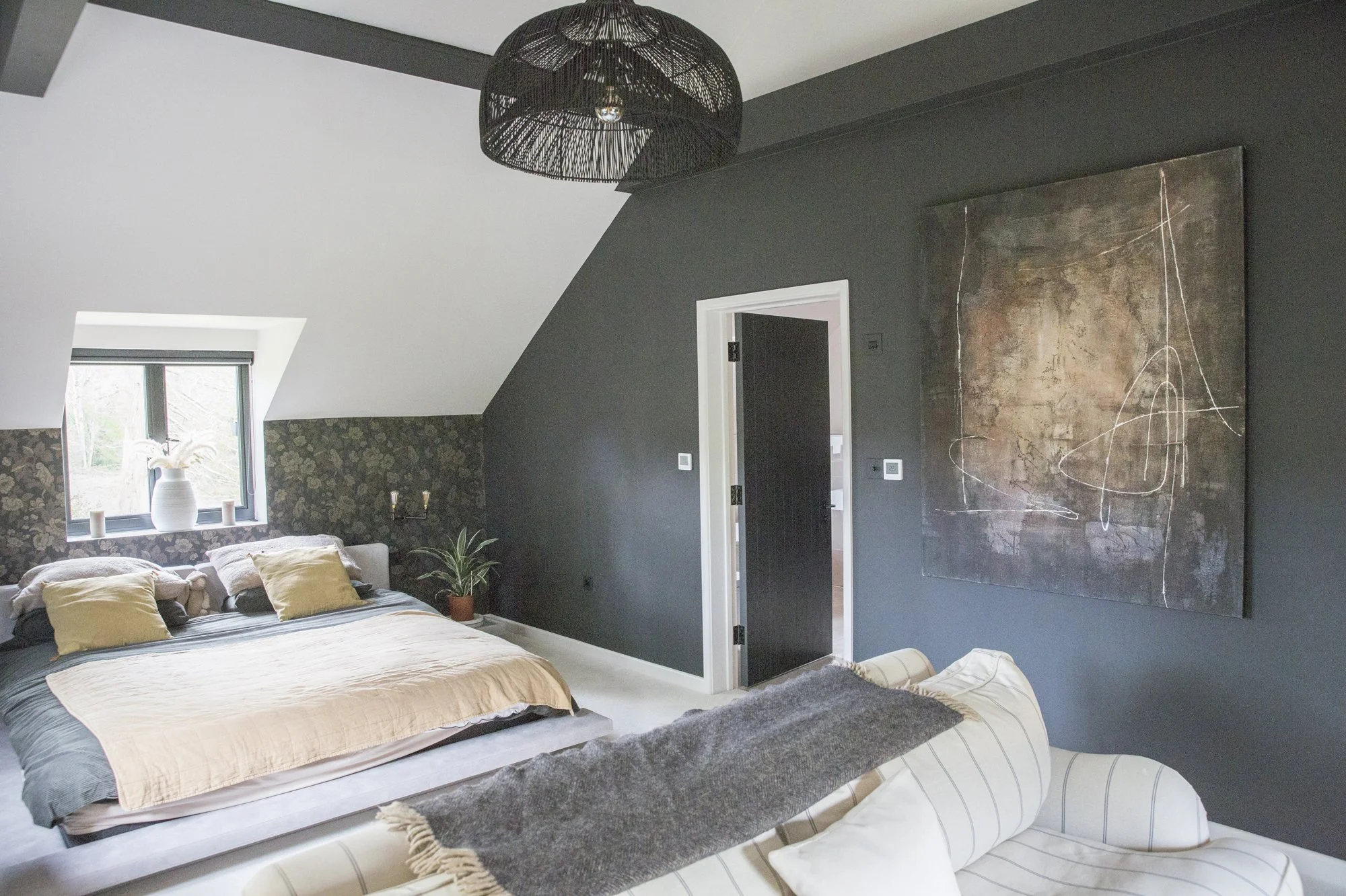 A bedroom with sloped ceilings, dark gray walls, a window with a white vase and decorative items, a beige bed with pillows, a large abstract painting, a small plant, and a black wire pendant light.