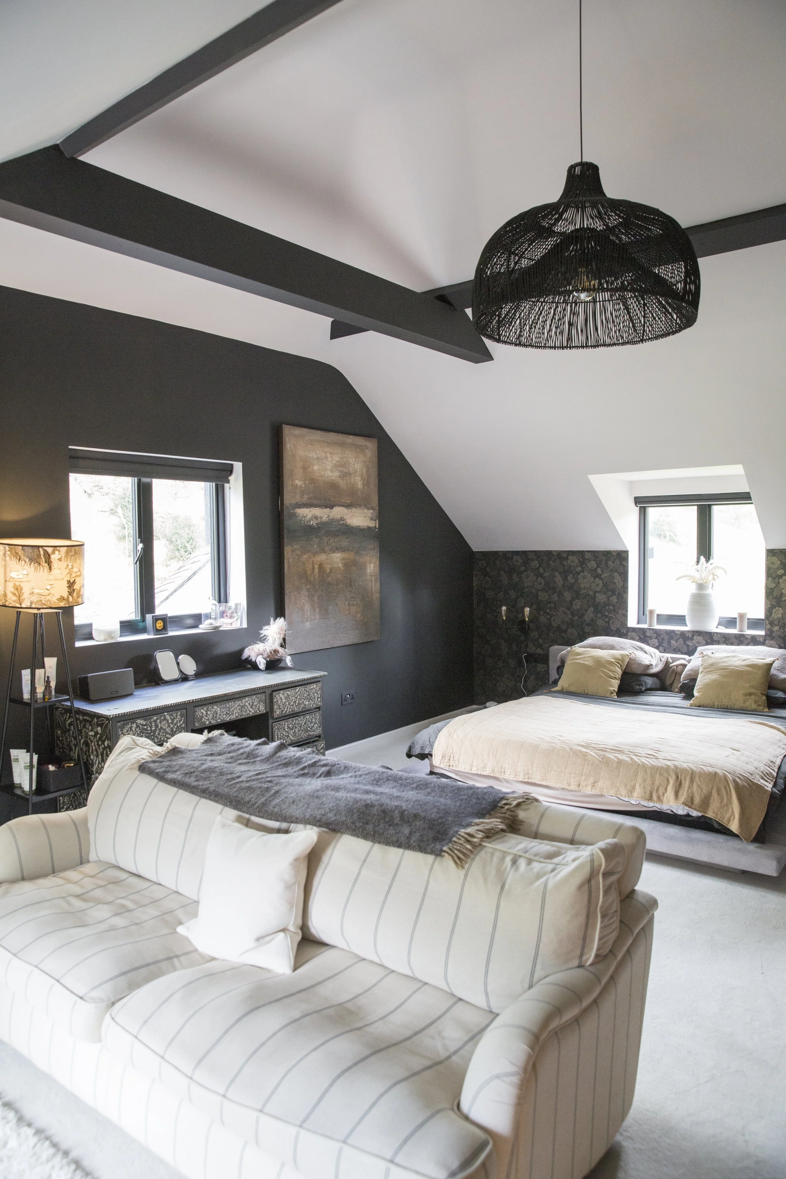 A cozy bedroom with dark walls, a large bed with beige bedding and pillows, a patterned sofa with a blanket, black pendant light hanging from the ceiling, two windows with blinds, and decorative items including a lamp and artwork.