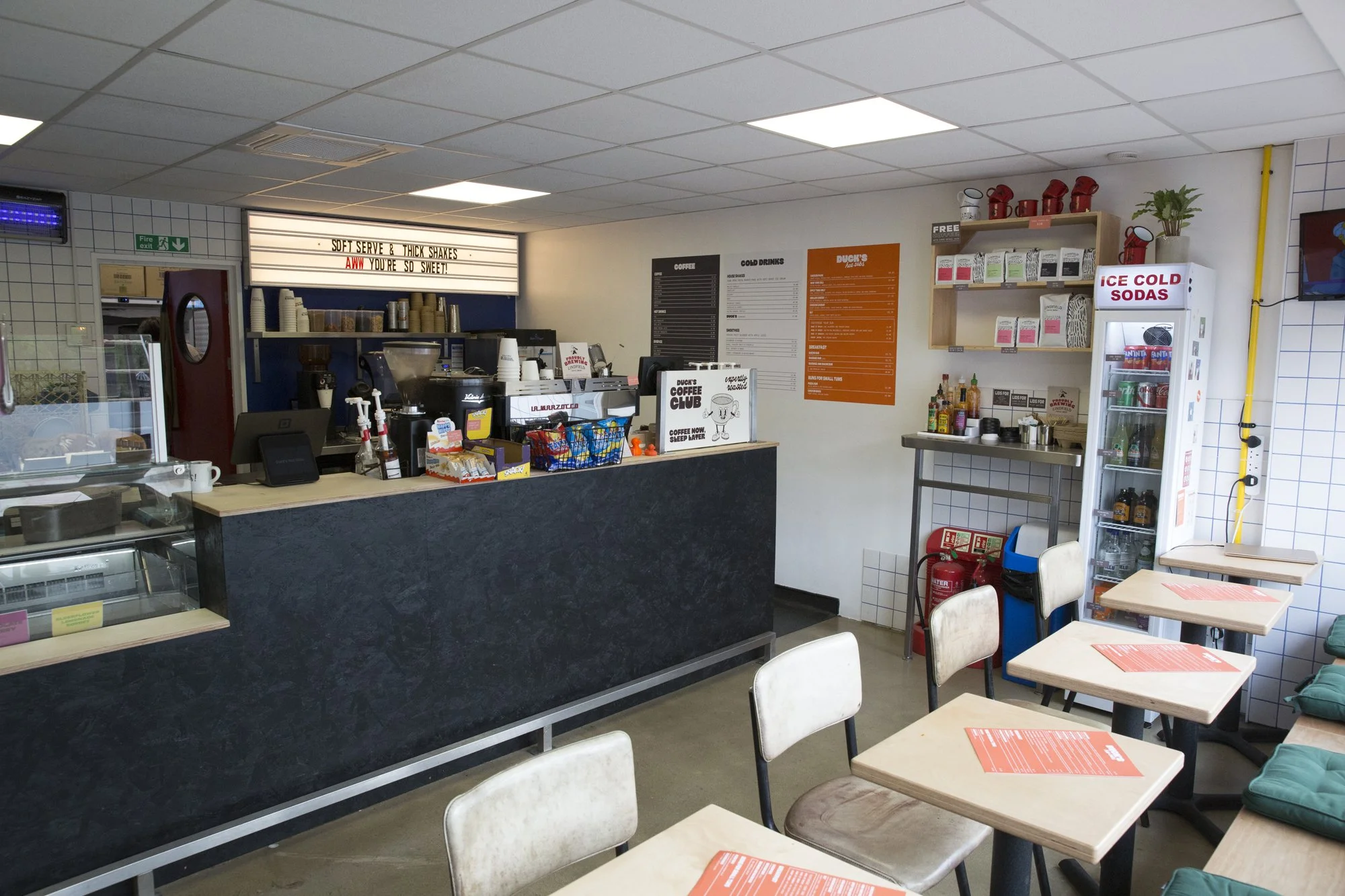 Interior of a small café with a counter, menu boards, and tables with chairs. The counter has a coffee machine, snack baskets, and paper cups. A sign above the counter reads: 'Soft serve & thick shakes. Aww you're so sweet!'. There's a white refriger