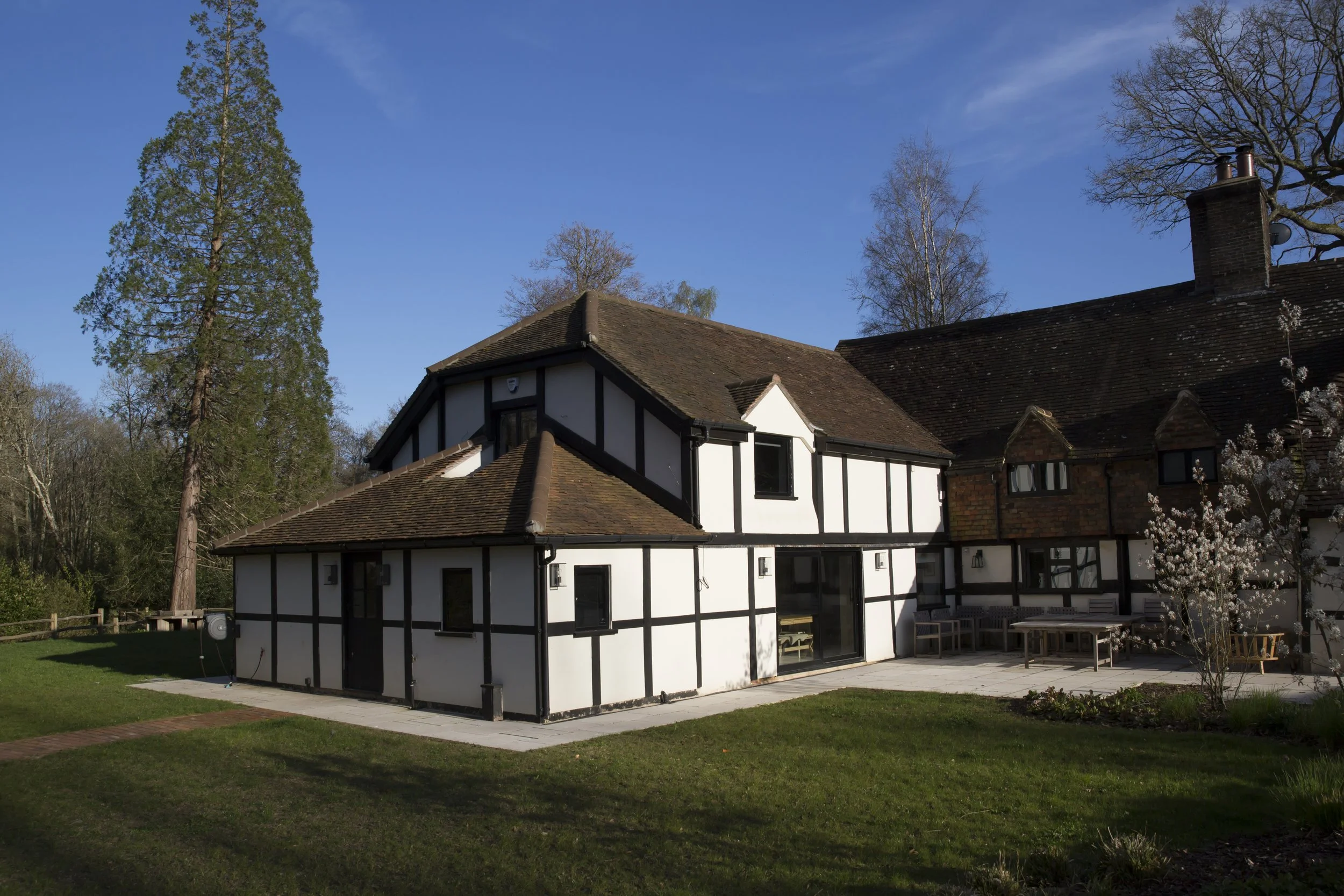 A house with black and white timber framing and a brown tiled roof, set against a backdrop of trees and a bright blue sky.