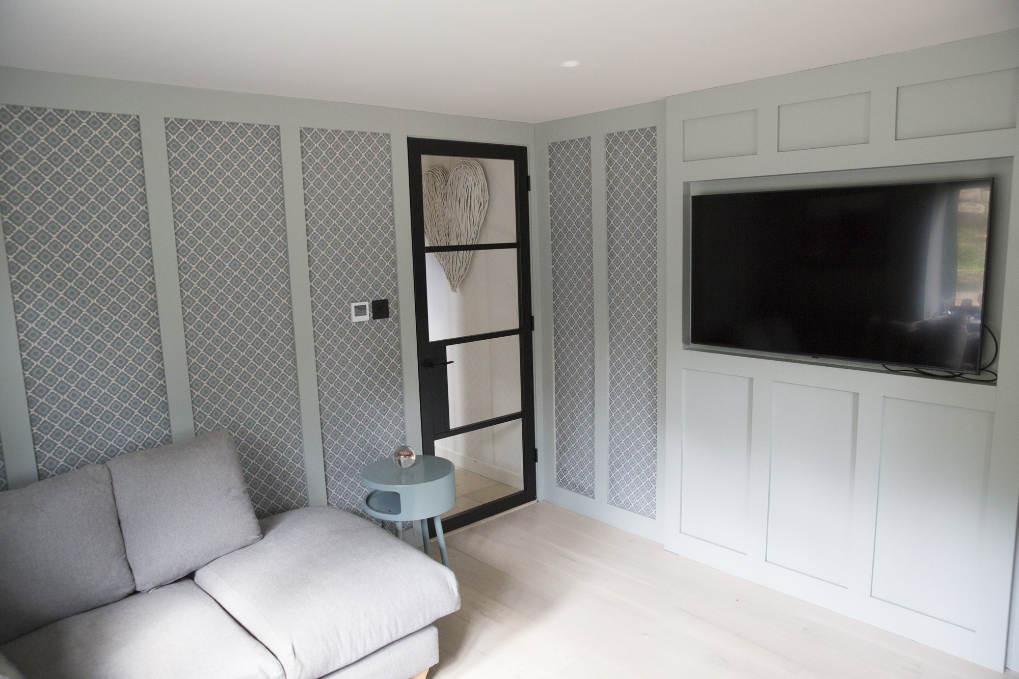 A living room with a light gray sofa, a small round blue side table with a candle, patterned wallpaper with gray and white design, a black door with a white heart-shaped wall hanging, and a mounted black flat-screen TV on white paneled wall.