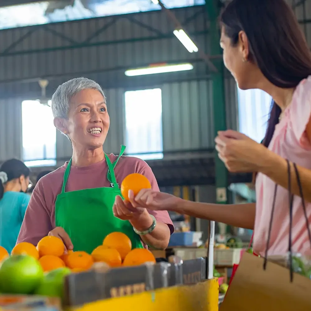 A woman in a pink shirt and green apron is smiling while handing an orange to a customer at a market stall with oranges and green apples, inside a market with high metal walls and fluorescent lighting.