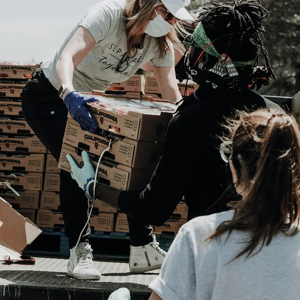 Two women are loading boxes of strawberries onto a truck, wearing masks and gloves.
