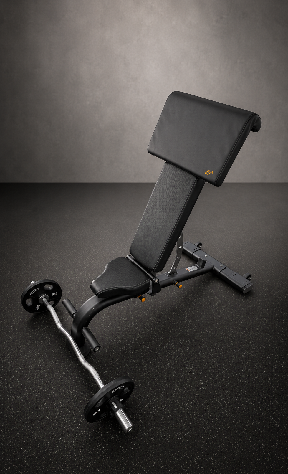 A black adjustable weight bench with a preacher curl pad attachment and a small weight plate with a barbell in the foreground on a speckled black gym floor.