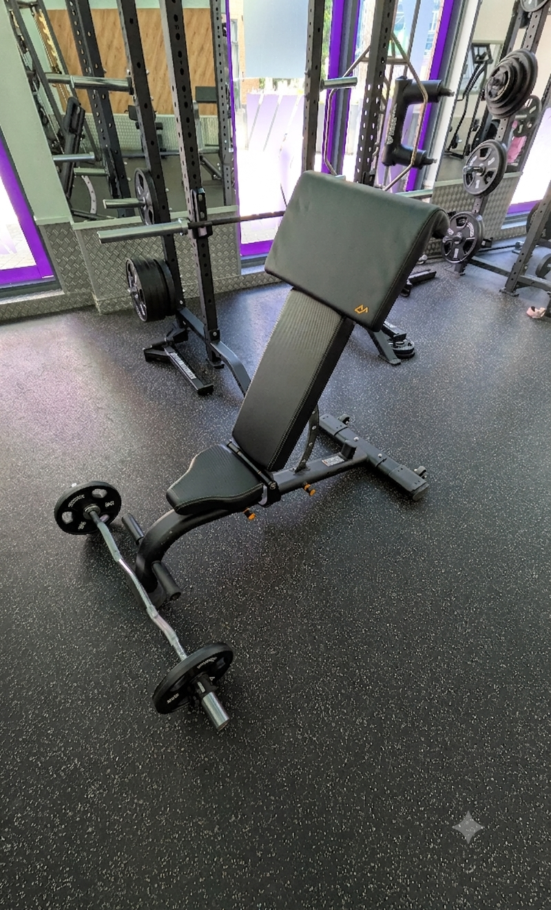 Adjustable workout bench with a barbell and weight plates on the floor in a gym.
