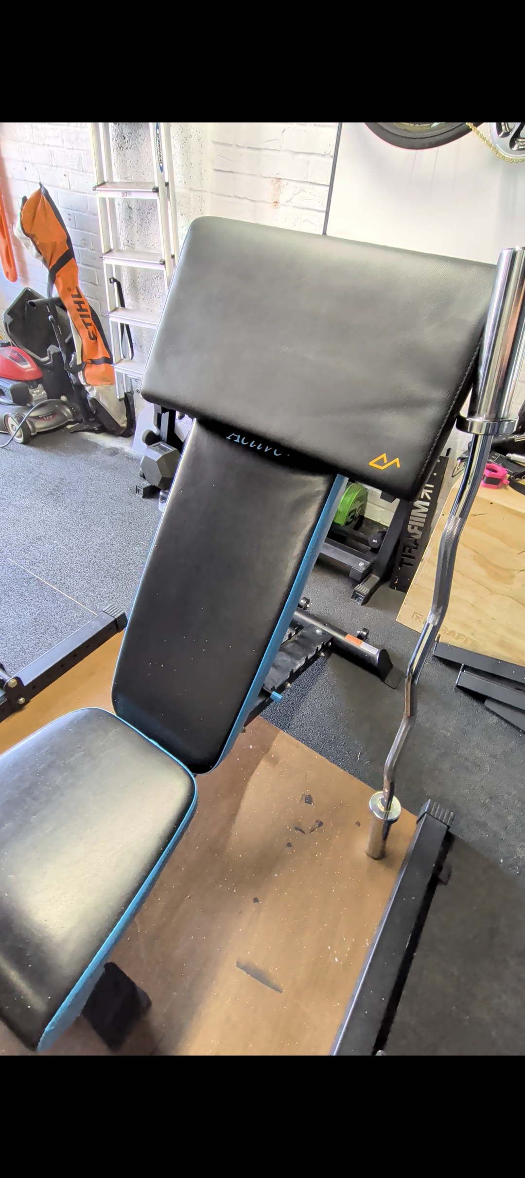 Adjustable workout bench with a preacher pad attached to it, in a gym with equipment and brick wall in background.