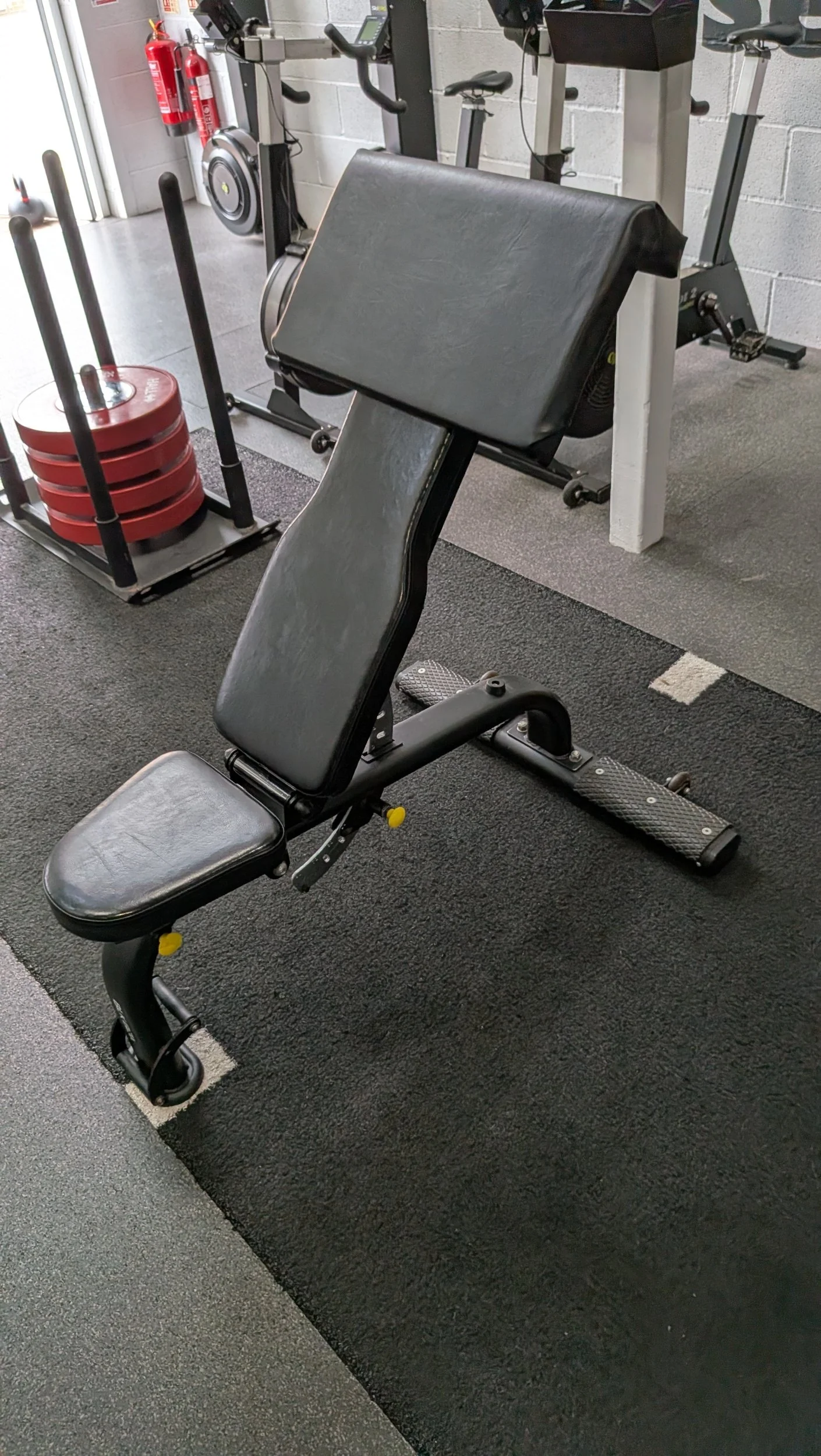 A workout gym with adjustable incline bench in the foreground with a preacher pad attached to it, a weight sled with red weights, and various cardio machines including an exercise bike and a treadmill, with red fire extinguishers mounted on the wall.