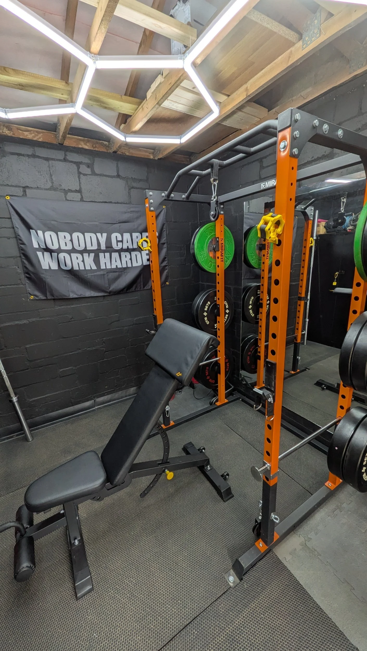 A home gym with a black brick wall, a black and orange power rack with weight plates, a black adjustable bench with a preacher pad at, gym mats, and a hexagonal LED light fixture on the ceiling. A banner on the wall reads, "NOBODY CARES WORK HARDER."