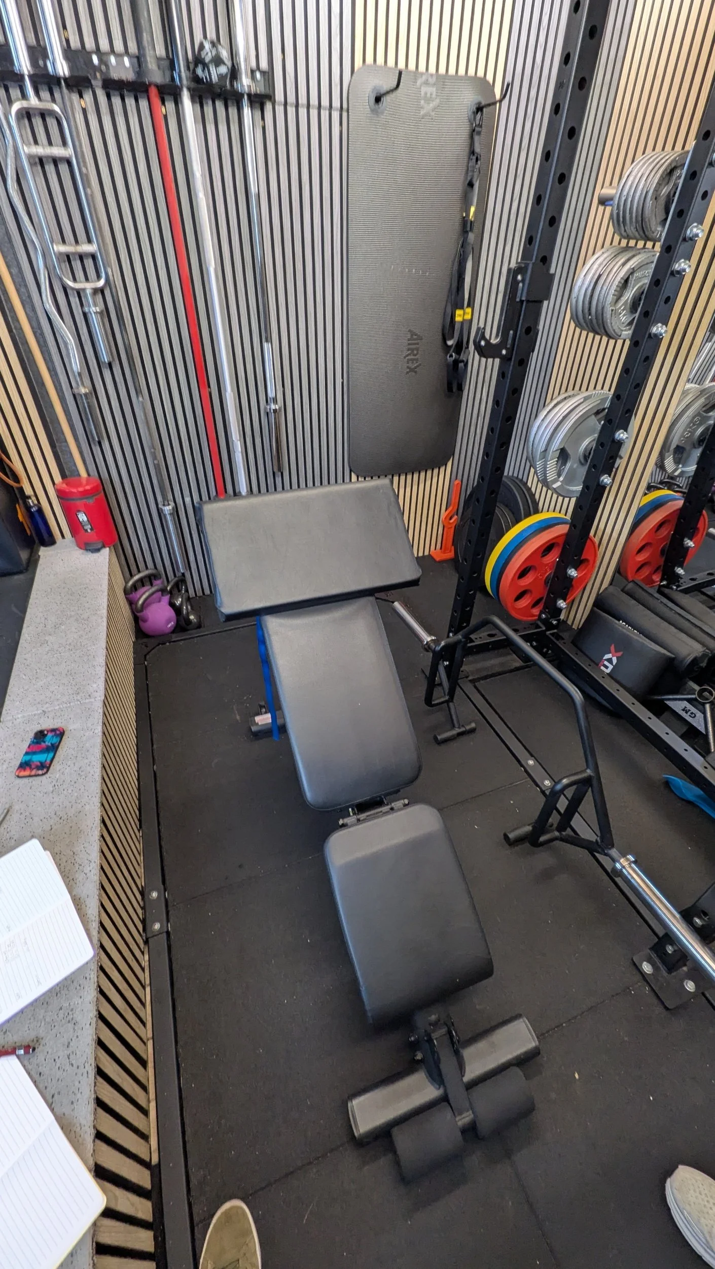 Indoor gym with various exercise equipment including a workout bench with preacher curl pad attached, dumbbells on a rack, weight plates, resistance bands, and a mat on the wall.