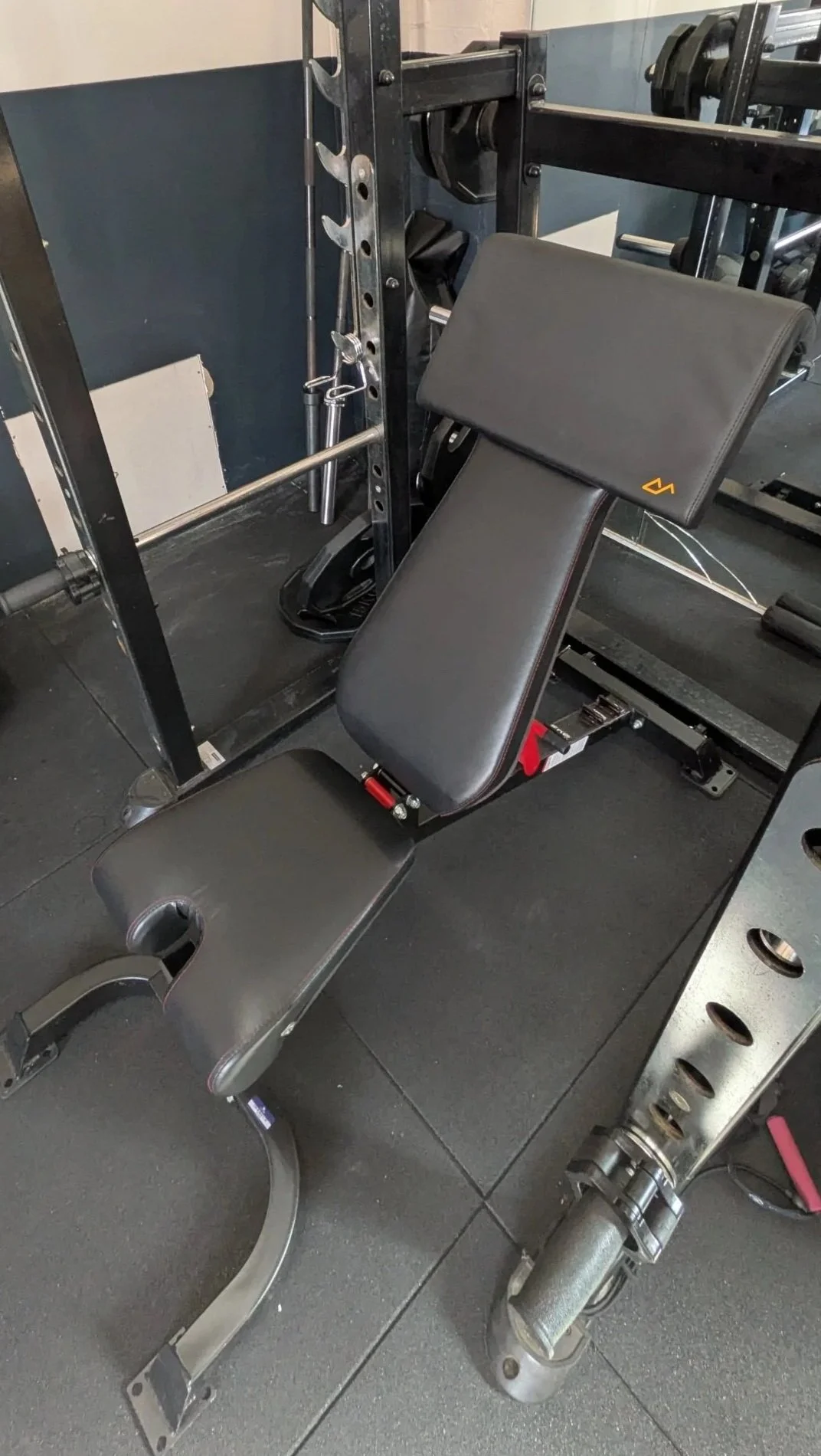 A black adjustable workout bench with a padded seat and preacher pad attached positioned in a gym near a metal power rack.