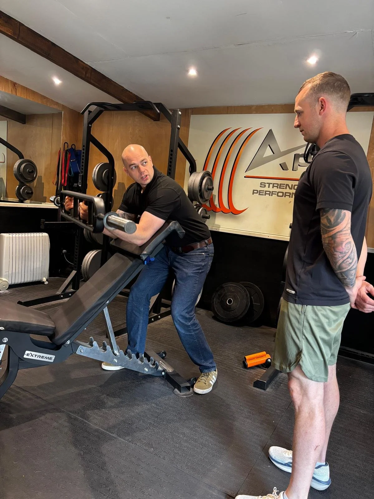 Two men in a gym, with one man using a preacher pad and the other man observing or coaching. The gym has various weights and equipment in the background, including a sign that says 'Strength Performance'.