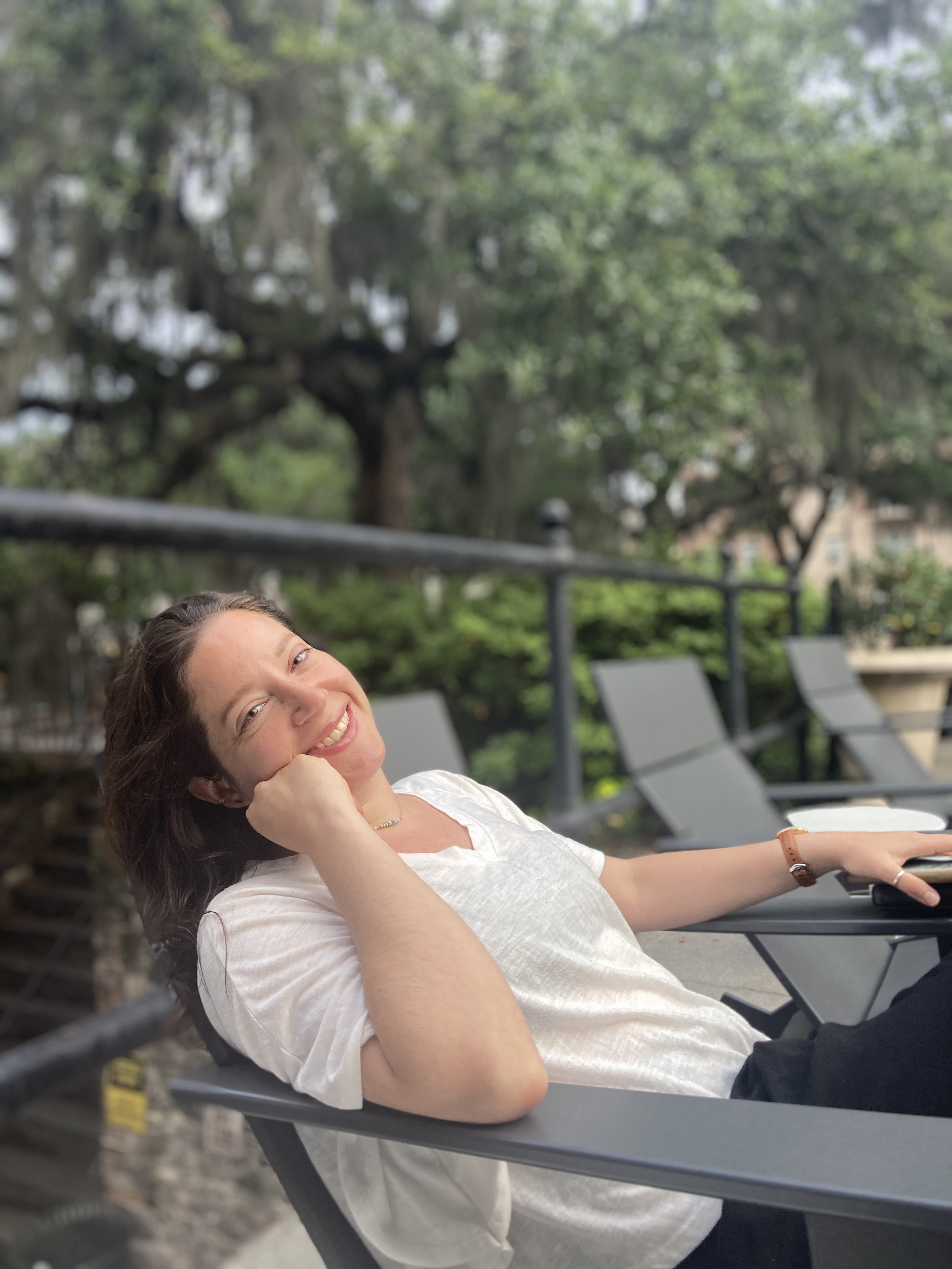 A woman with long brown hair, smiling, lying on a lounge chair outdoors with trees and greenery in the background.