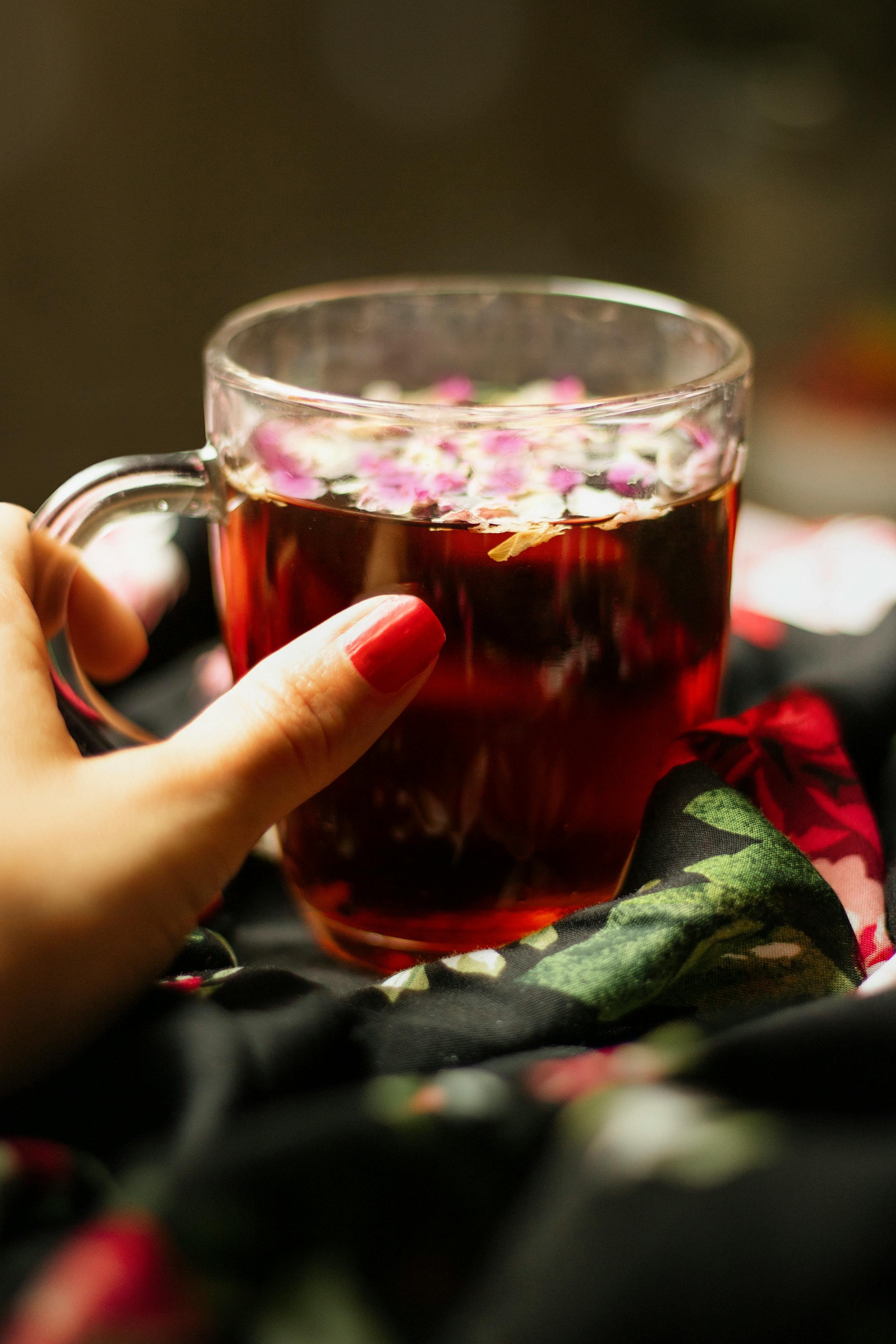 A close-up of a clear glass mug filled with dark red tea, held by a hand with red-painted nails, against a dark background with out-of-focus floral fabric.