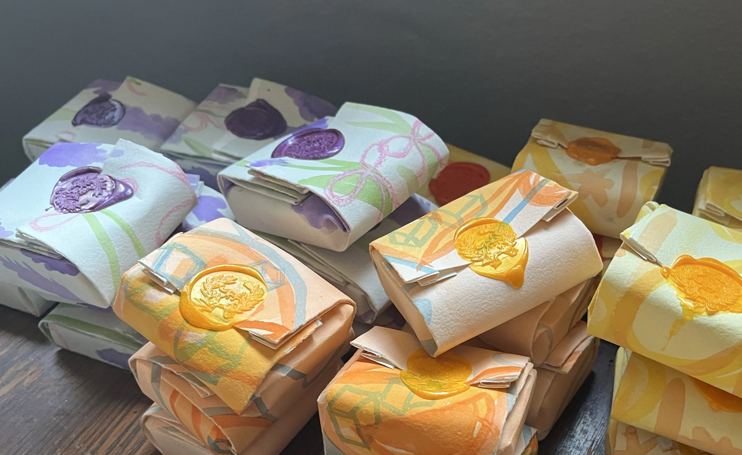 A collection of handmade soap bars wrapped in colorful, patterned paper, each topped with a wax seal in various shades of purple, orange, yellow, and red.