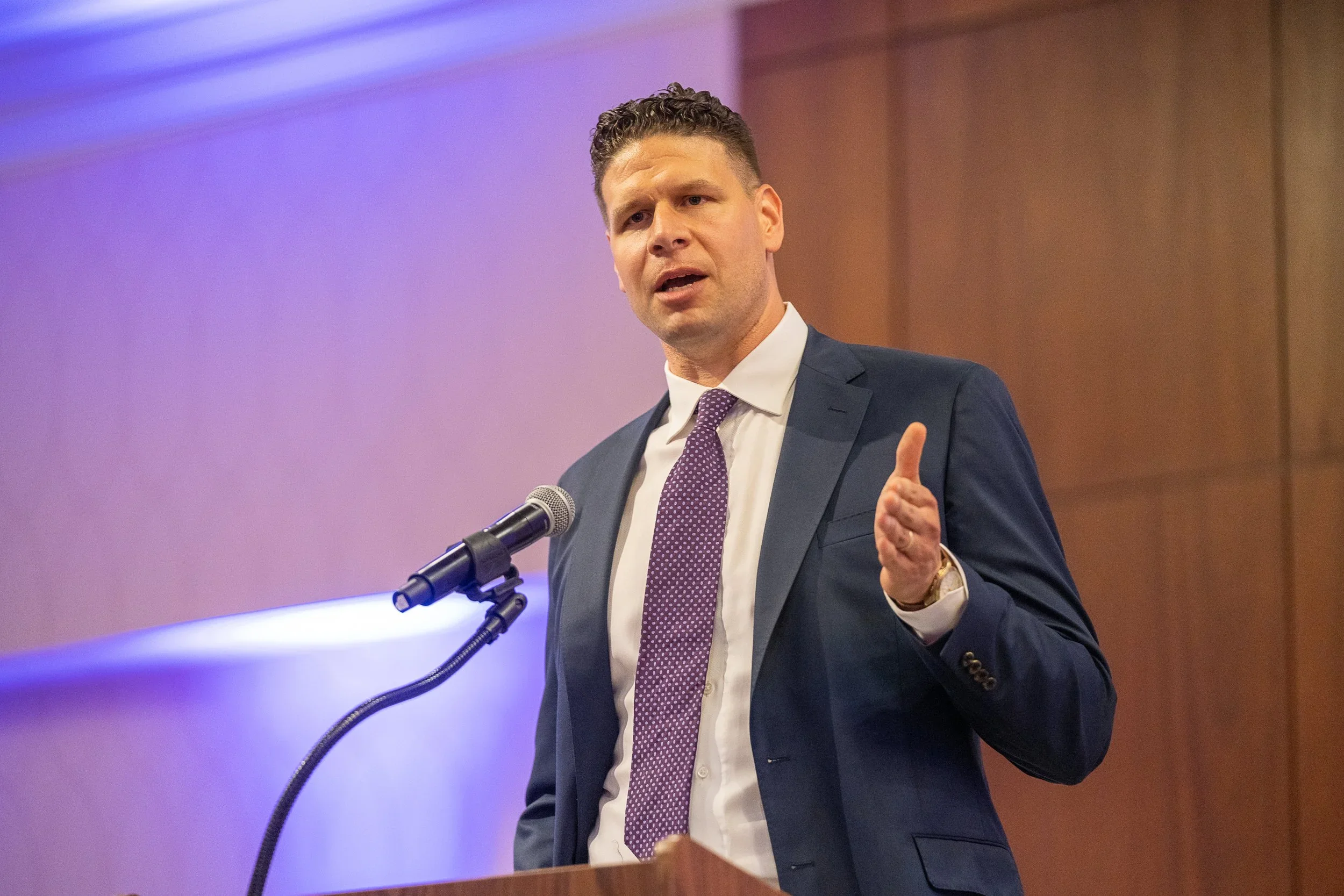 A man in a dark suit and purple tie speaking at a podium with a microphone, gesturing with his right hand, in a conference or presentation setting.