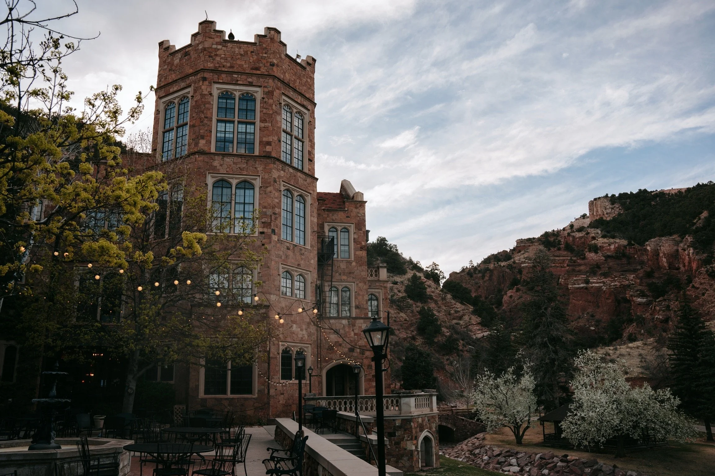 A historic stone building with tall windows and a tower, set against a mountainous landscape, with trees and outdoor seating in the foreground.