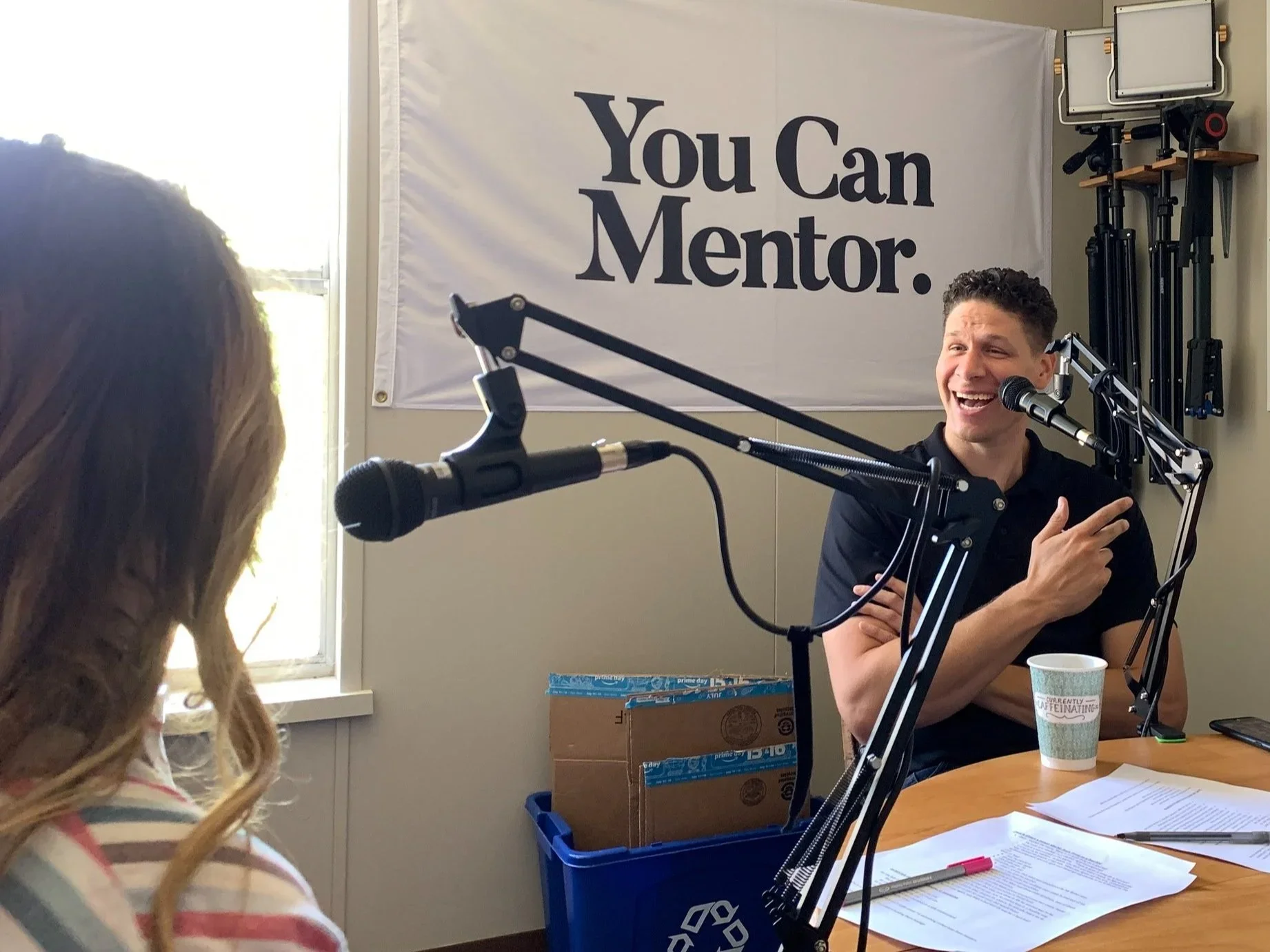 A man and a young girl appear in a podcast or interview setting, with the man speaking into a microphone and a window in the background. A large banner on the wall reads, "You Can Mentor." The table has papers, a pen, and a cup, with boxes and equipment visible behind them.