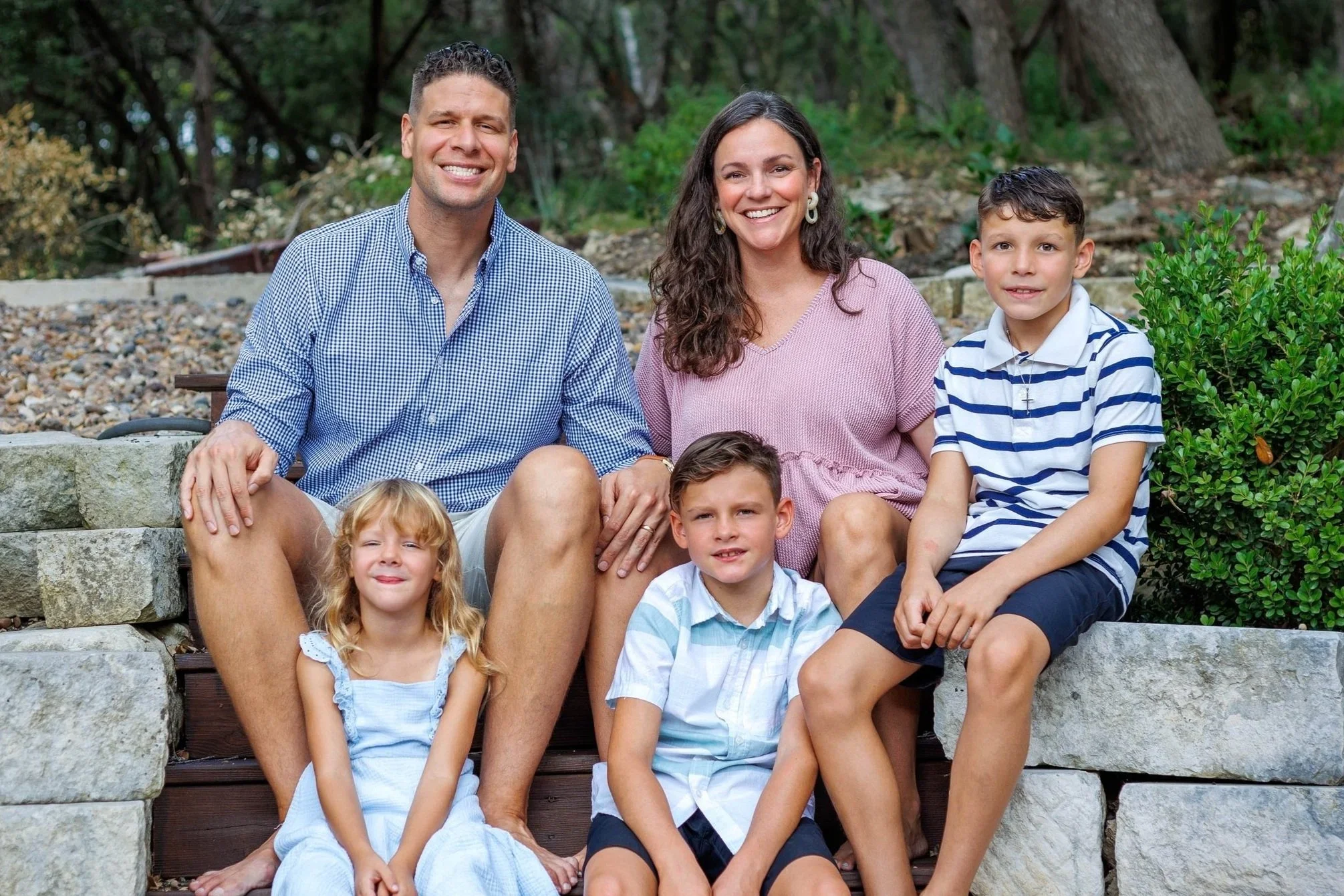 A family of seven sitting on stone steps outdoors, smiling at the camera. The family includes two adults (a man and a woman) and five children, with a lush green background of trees and shrubbery.