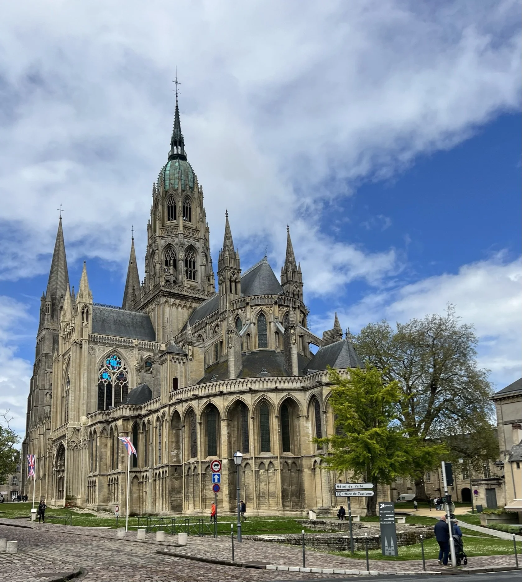 A Gothic cathedral with tall spires and detailed architecture, situated in an urban area with cobblestone streets and people walking nearby, trees and signs in the foreground, and a partly cloudy sky above.