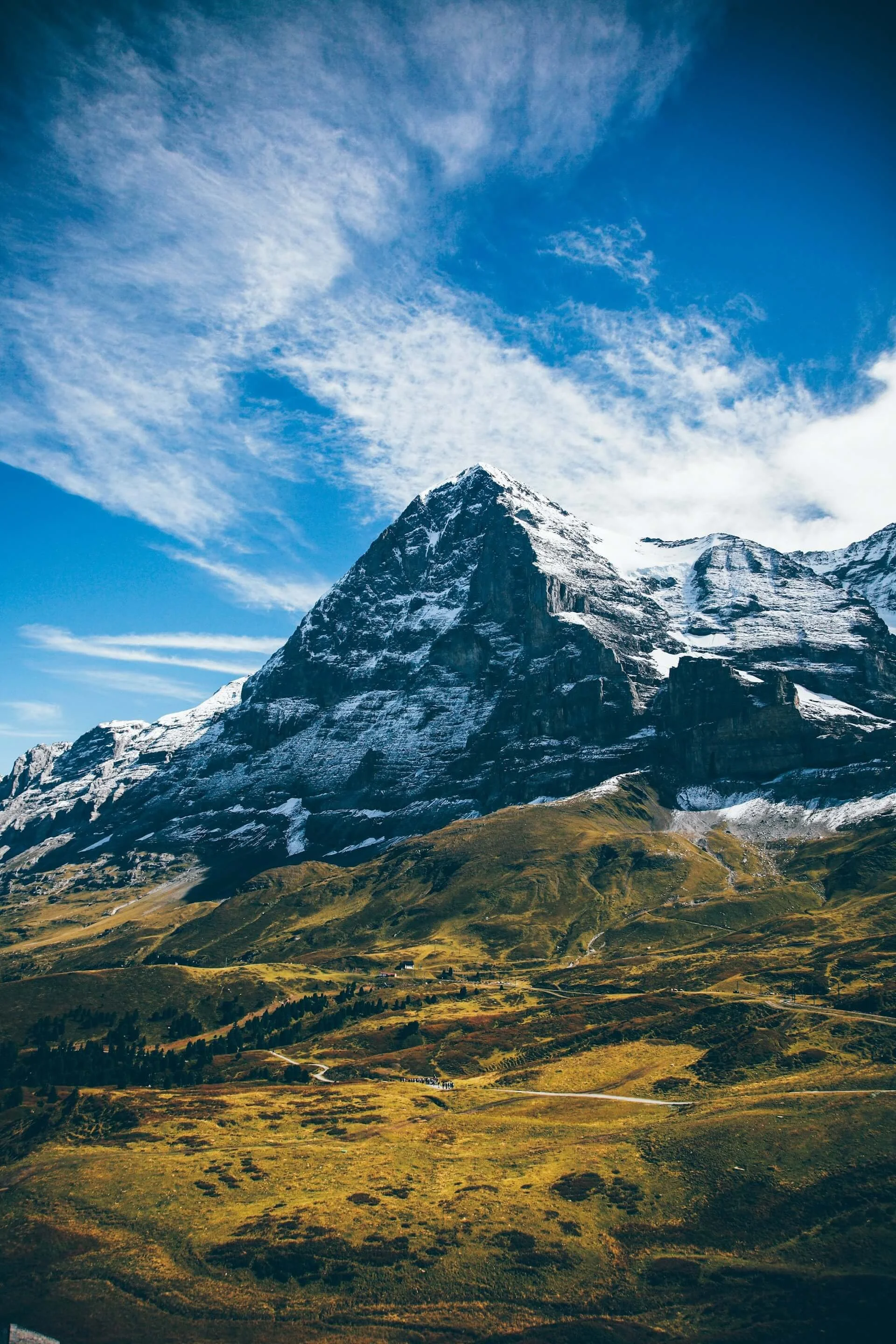 Snow-capped mountain in a clear blue sky with scattered clouds, green rolling hills in the foreground.
