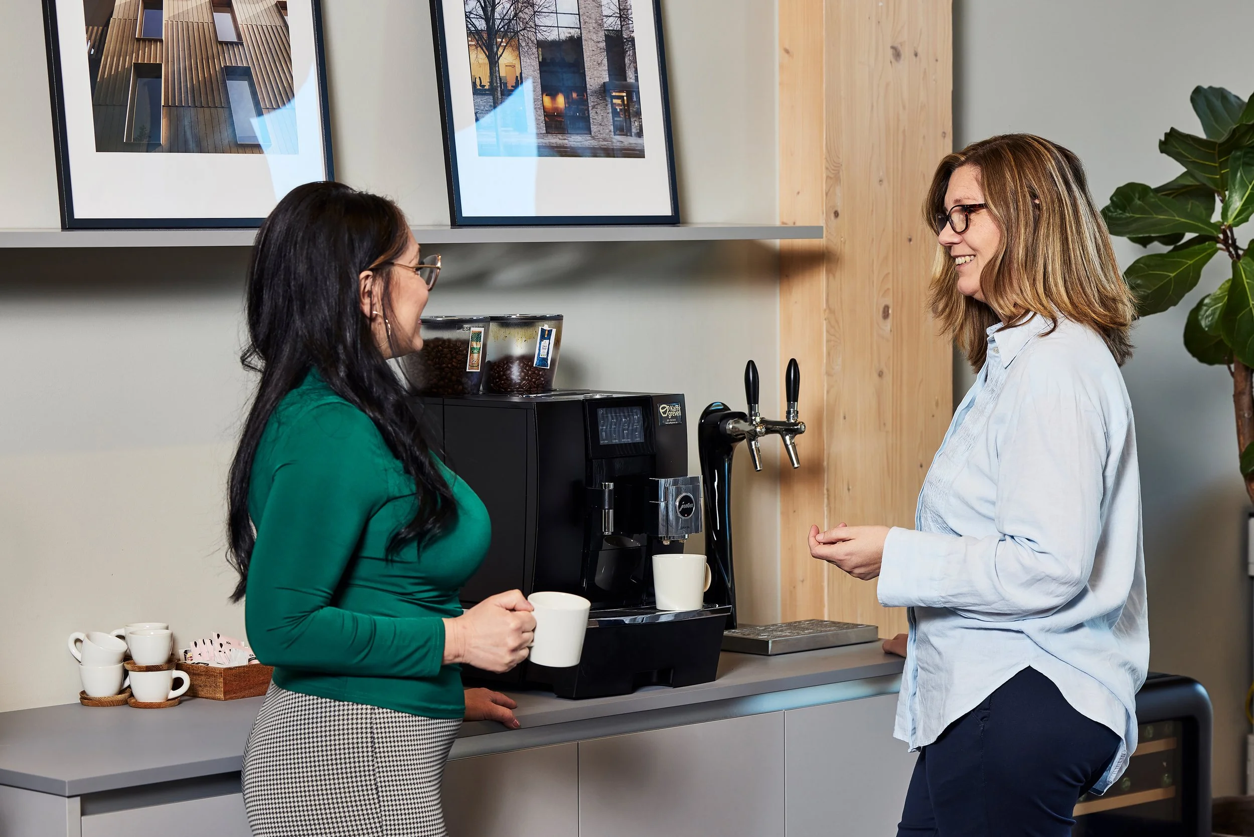 Två kvinnor pratar vid ett kaffemaskin i ett kök, den ena håller en kaffemugg, det finns fler muggar och kaffekapslar på bänken.