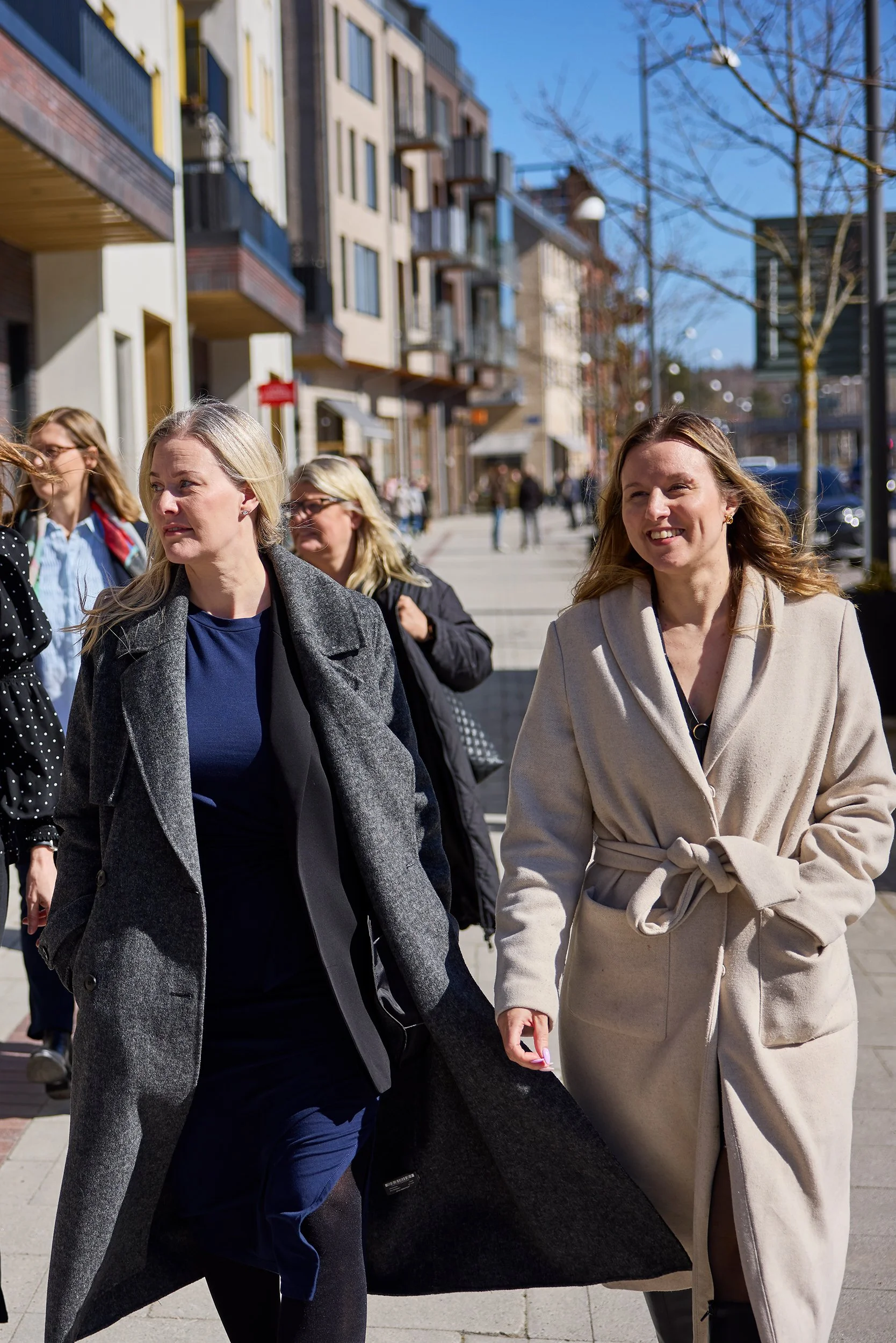 Flera kvinnor som går på en trottoar i stadsmiljö, med moderna byggnader och träd med späda grenar i bakgrunden, under en klar dag.