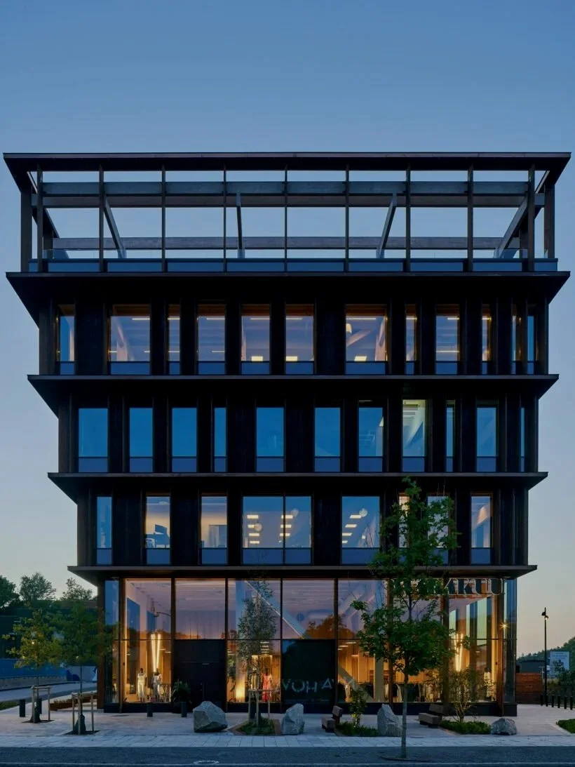 Modern kontorsbyggnad med stora glasfönster och flera våningar, omgiven av träd och sten.
