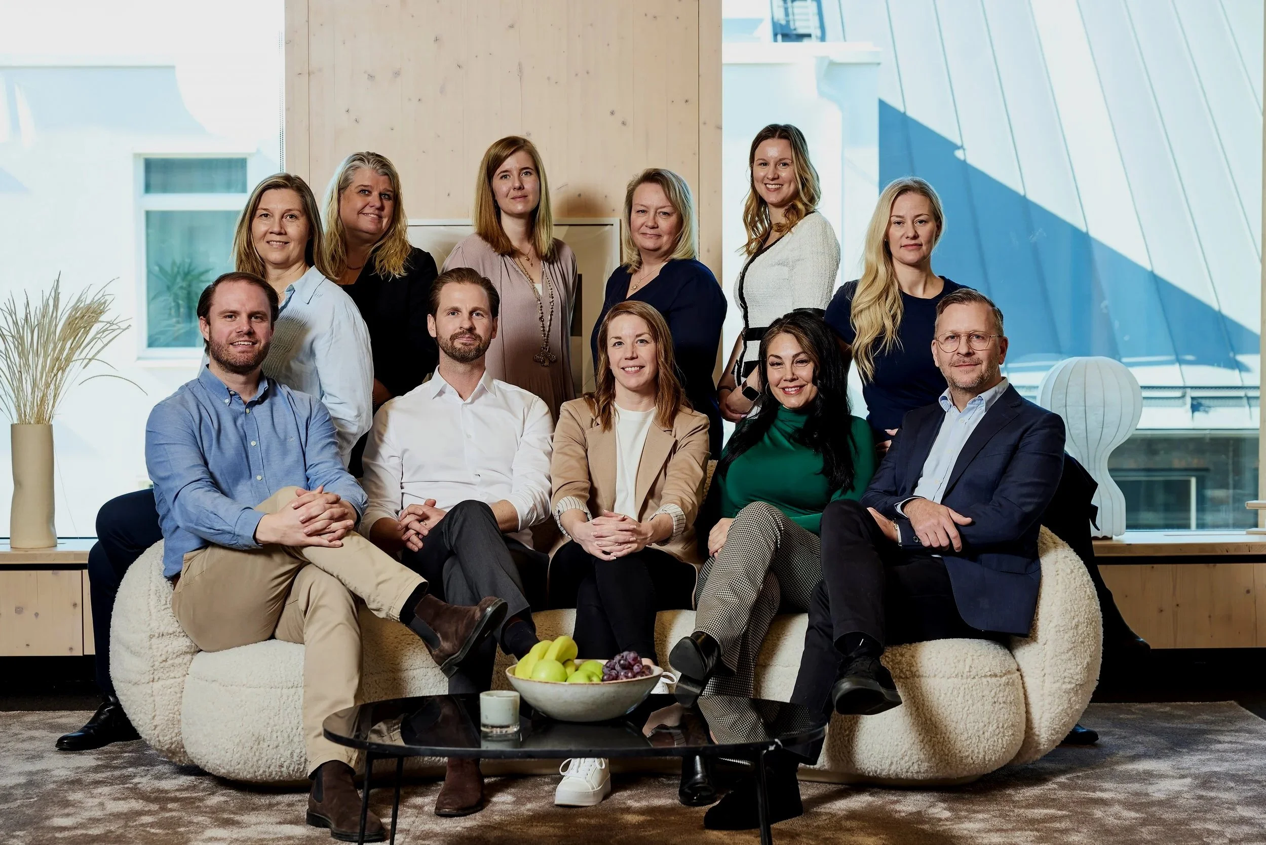 En grupp människor som sitter och står för ett fotografi i ett modernt kontorsutrymme med stora fönster och stadssiluett i bakgrunden. Det finns en soffa, ett svart runt soffbord med en skål frukt och en ljus vas med växt.