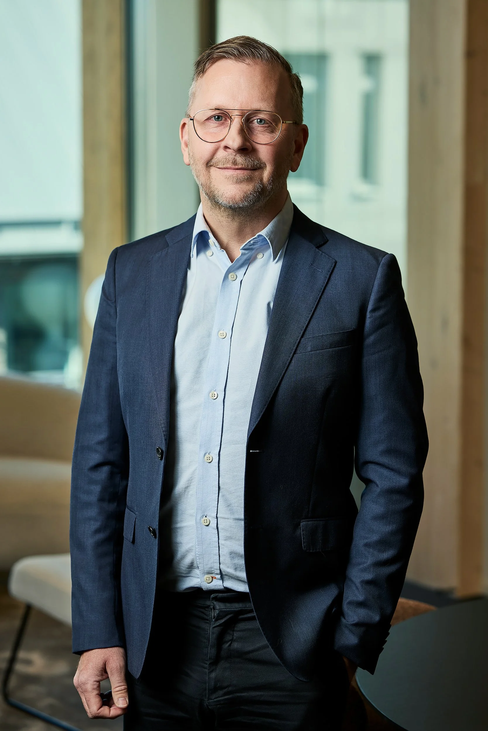 A middle-aged man with short light brown hair, glasses, and a beard, dressed in a navy blue suit jacket and light blue shirt, standing indoors near a window with a view of a building.