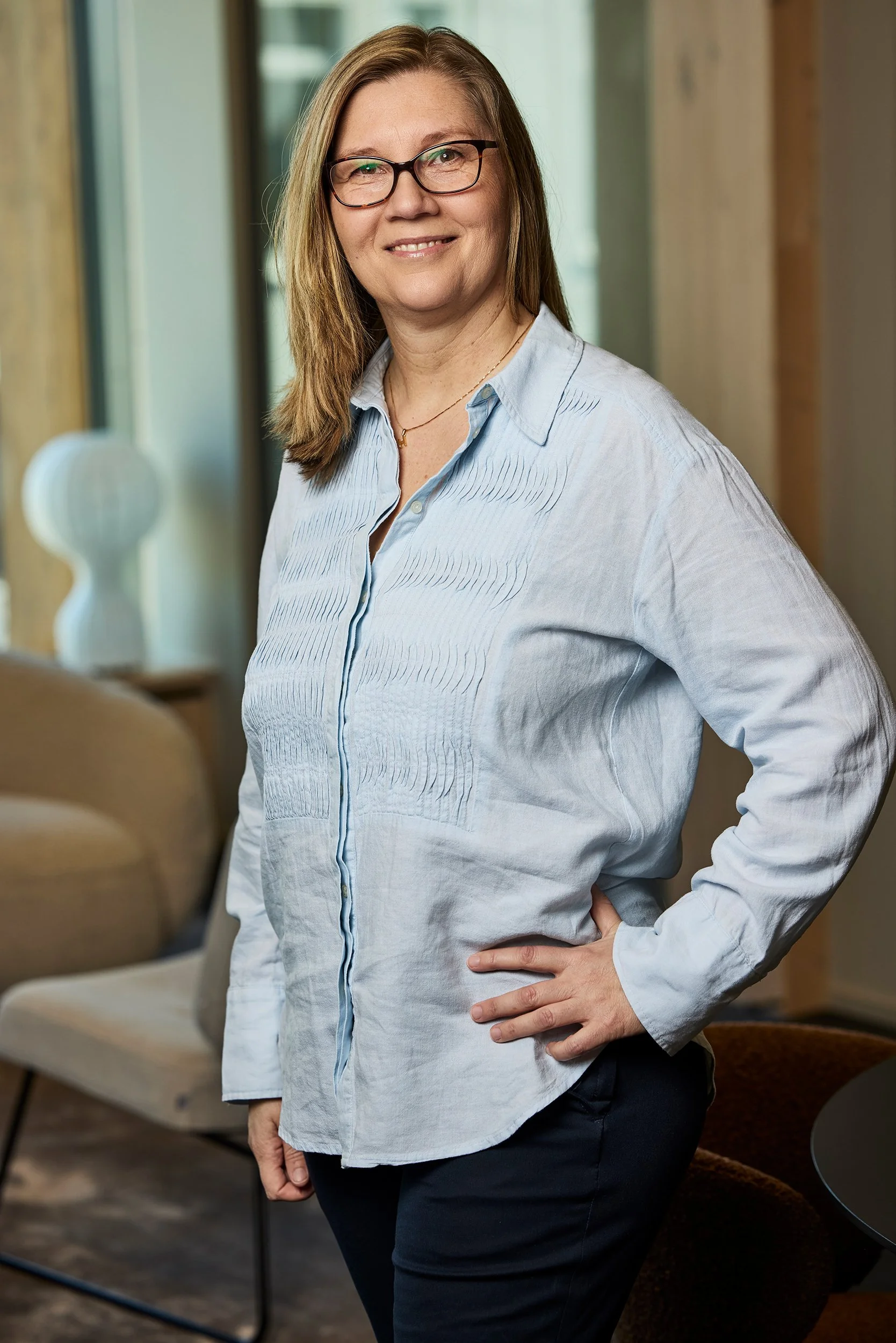 A woman with glasses and brown hair, wearing a light blue blouse and dark pants, standing in a modern indoor setting with her hand on her hip.