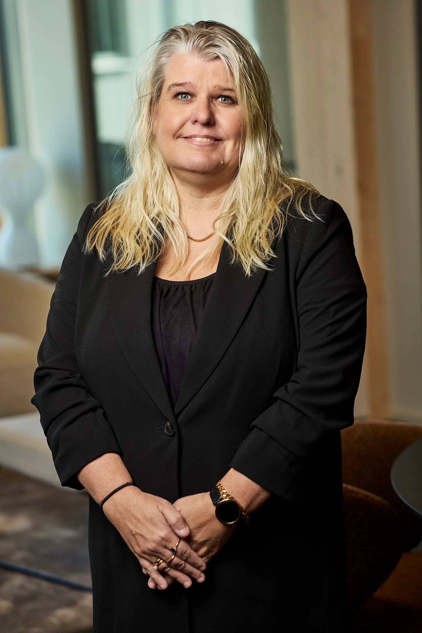 A woman with blonde, wavy hair smiles while standing in a modern office. She is wearing a black blazer, black top, and accessorized with a gold necklace, watch, and rings. The background features large windows and contemporary furniture.
