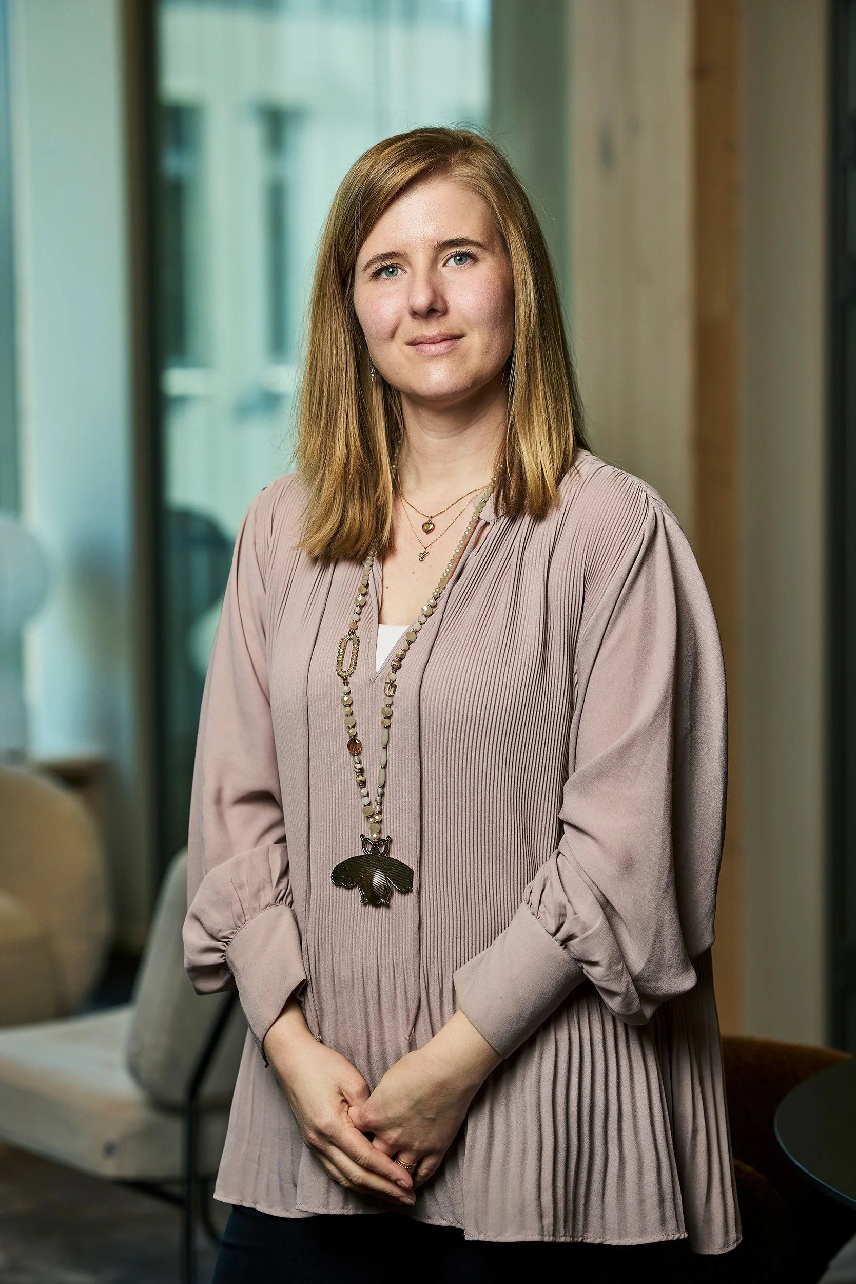A woman with shoulder-length light brown hair and blue eyes standing indoors, wearing a light mauve pleated blouse with long sleeves and layered necklaces, one with a large butterfly pendant.