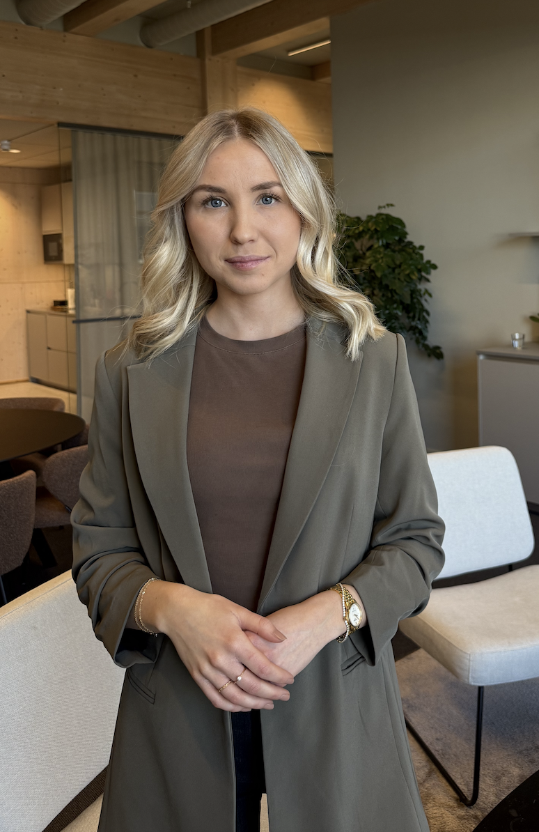 A young woman with blonde hair wearing a brown top and an olive green blazer, standing in a modern, well-lit indoor space with wooden ceiling beams and indoor plants.