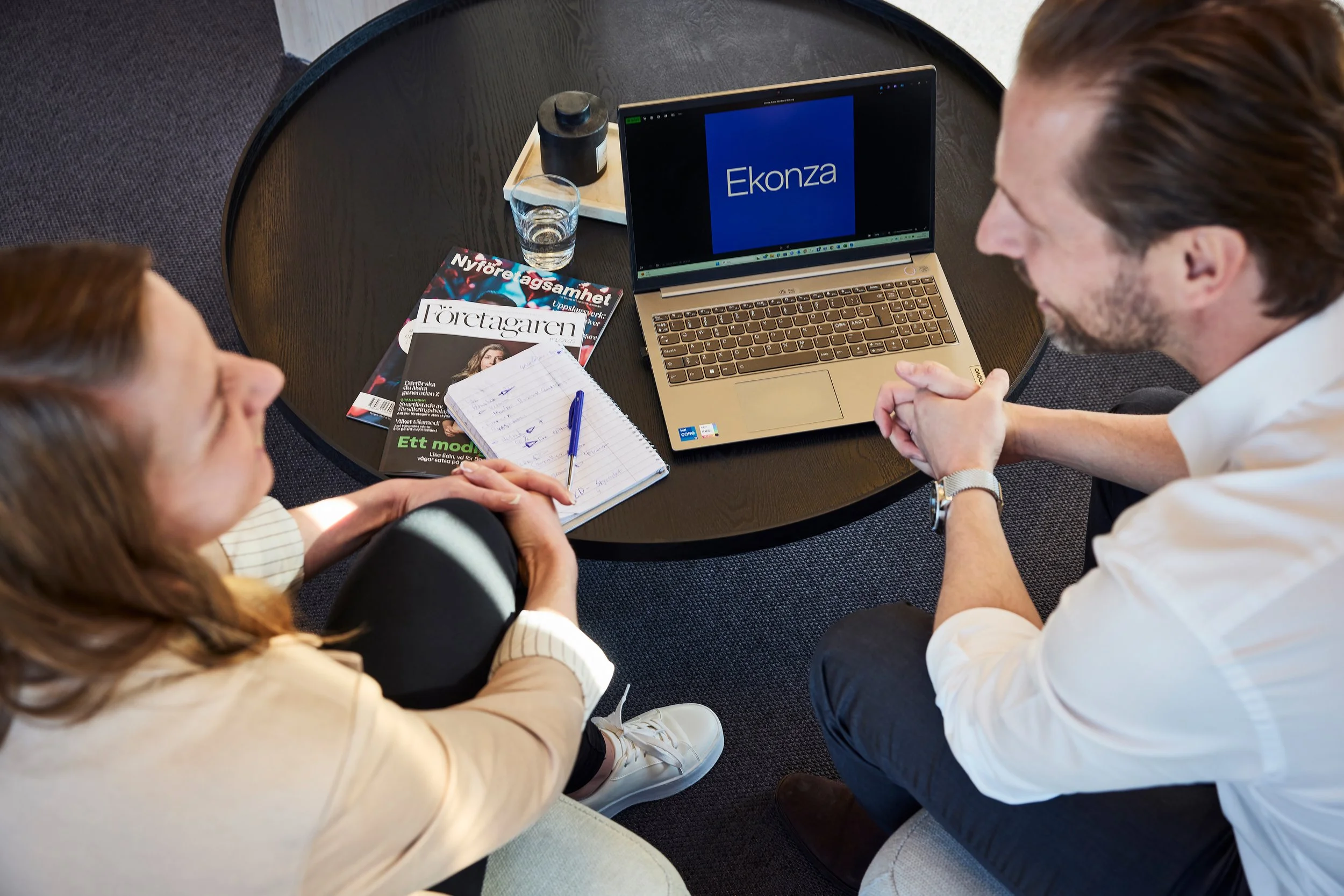 Two people sitting at a round table in a meeting or discussion, with a laptop displaying the word 'Ekonza', magazines, a glass of water, a notepad, and a pen on the table.