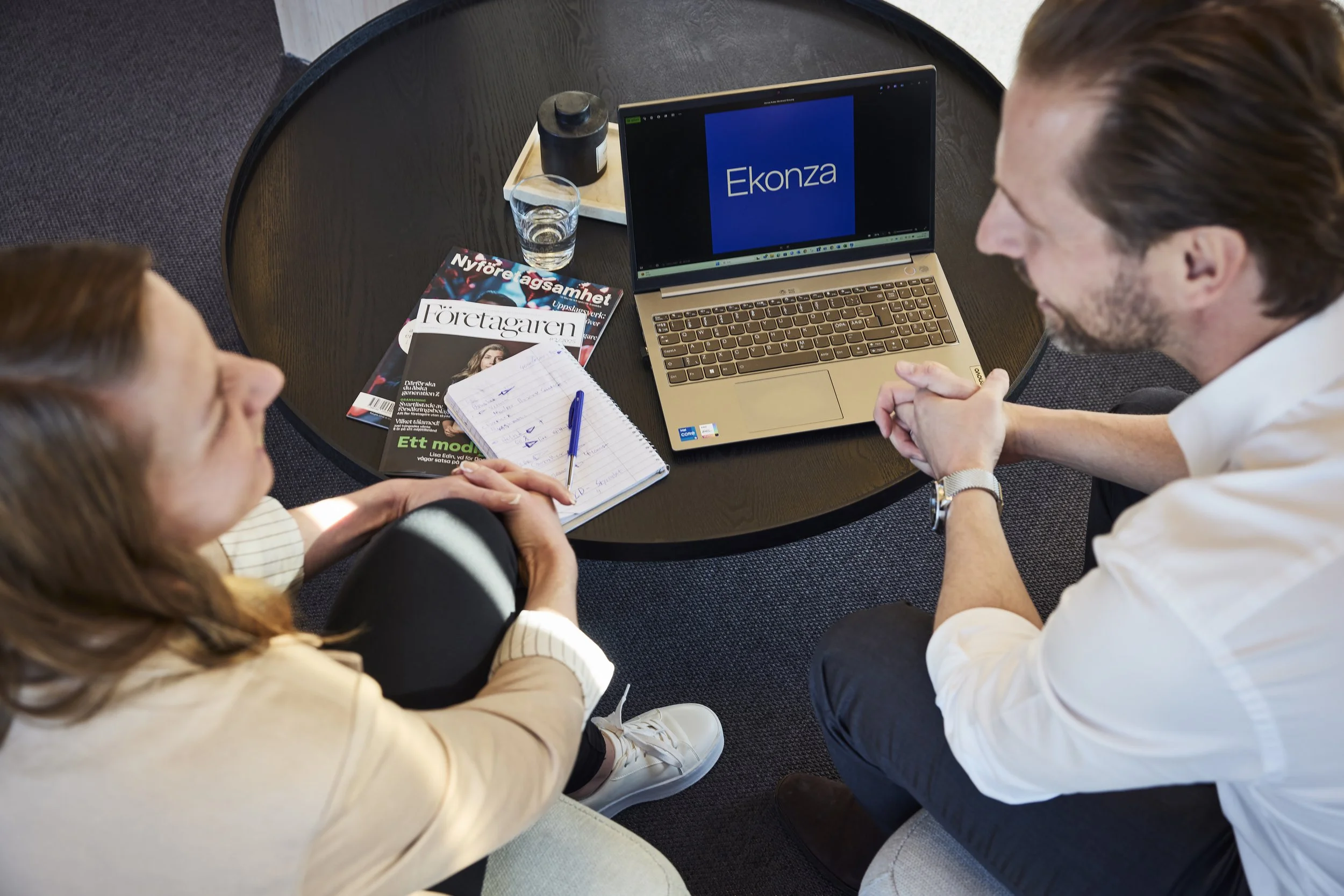 Two people sitting at a round table engaging in a business discussion, with a laptop displaying 'Ekonza,' magazines, a notepad, and a glass of water on the table.
