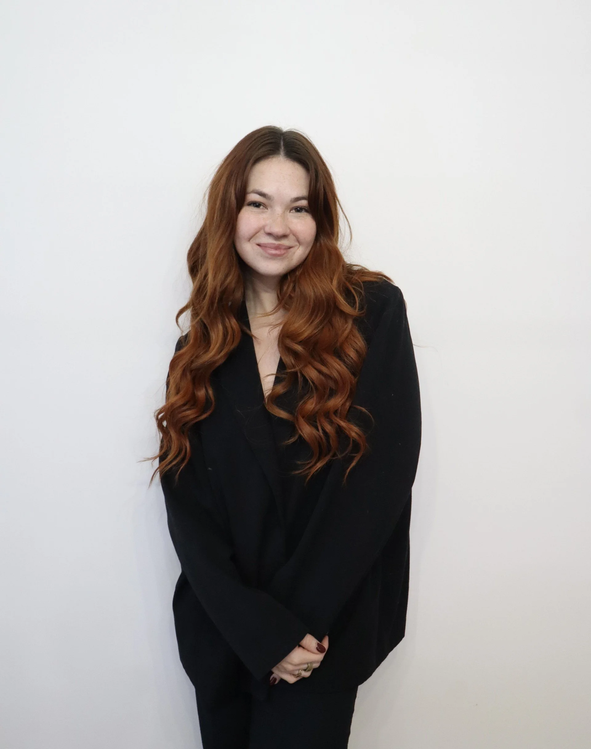 A woman with long, wavy red hair wearing a black blazer, standing against a plain white wall and smiling softly.