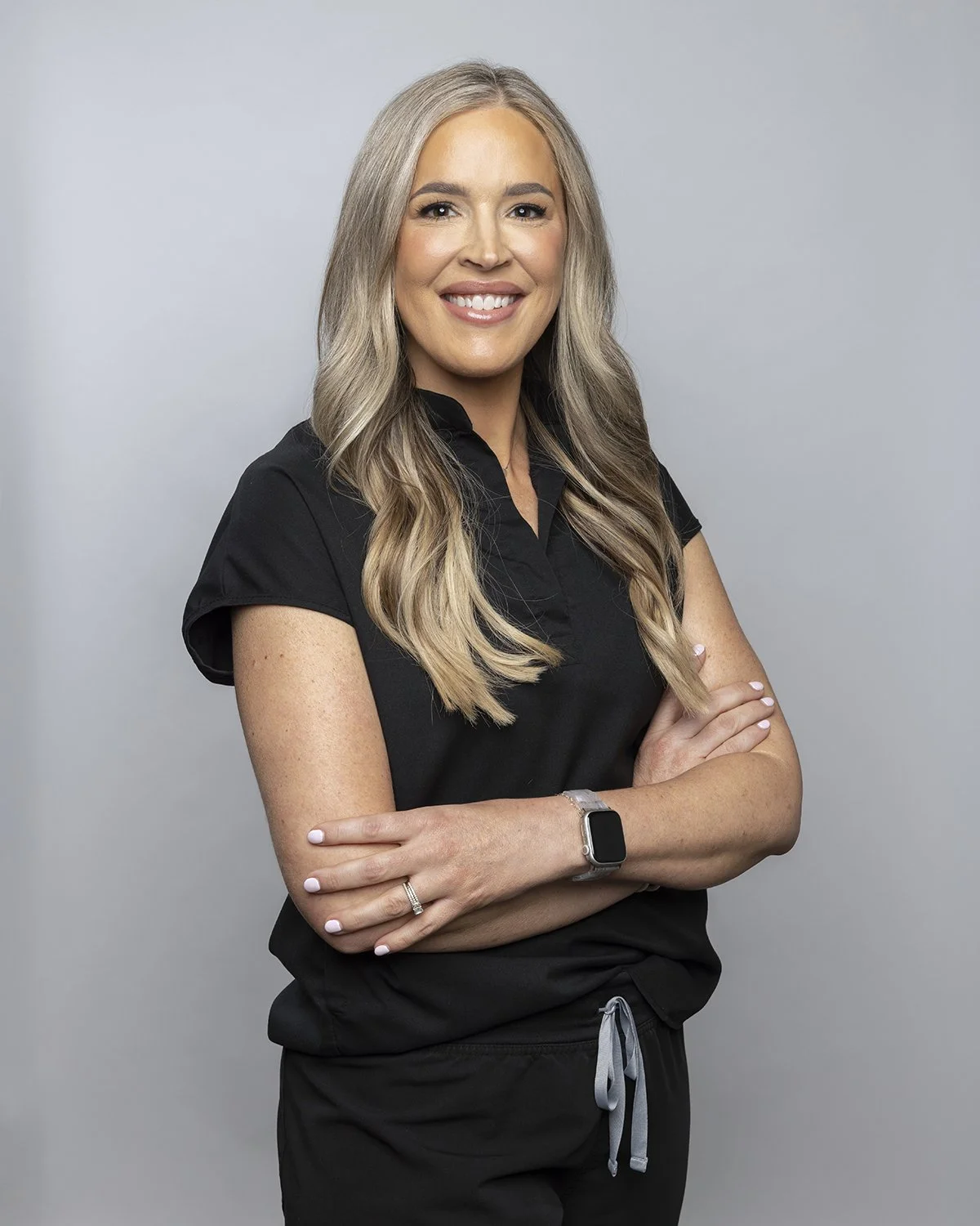 A smiling woman with long, wavy blonde hair wearing a black short-sleeve top, black scrubs, a smartwatch, and a ring, standing with arms crossed against a plain light gray background. Alaina Gray Aesthetics | Botox Springfield MO