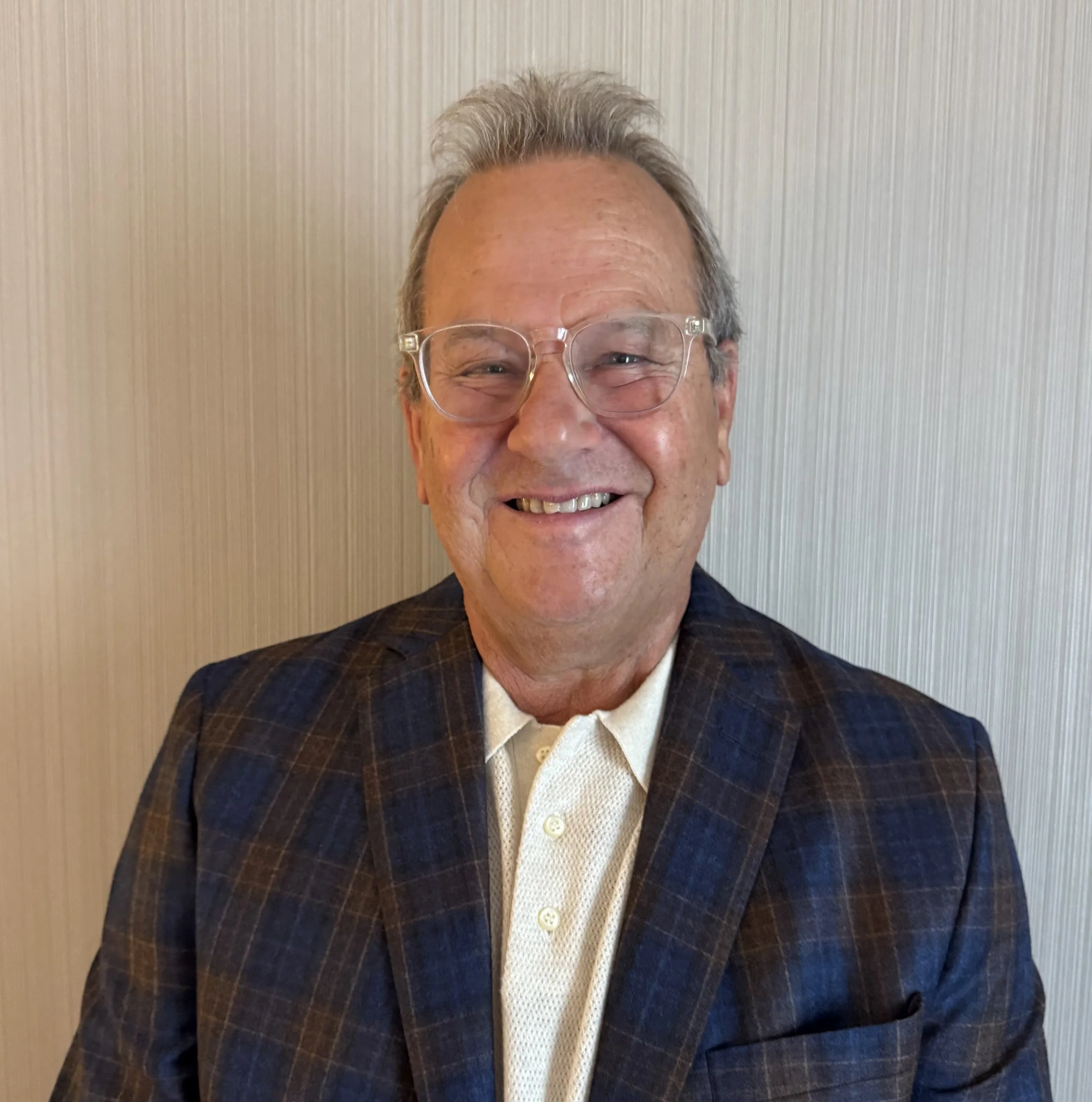 Close-up of a smiling elderly man with glasses, wearing a checkered blazer and a white shirt, standing against a light-colored textured wall. Alaina Gray Aesthetics | Botox Springfield MO
