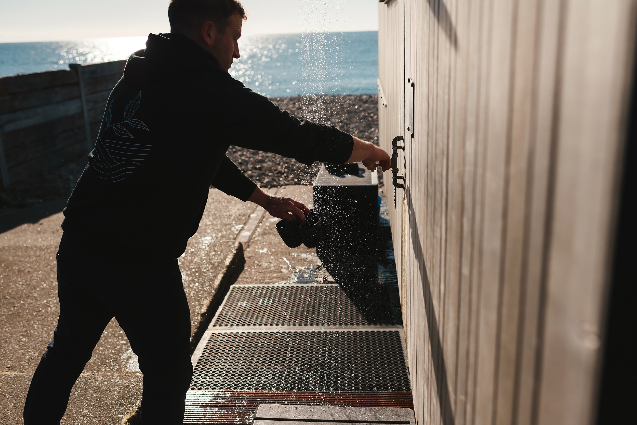 A man standing outdoors near the ocean, reaching into a sink or faucet, with water splashing and the sun reflecting off the water.