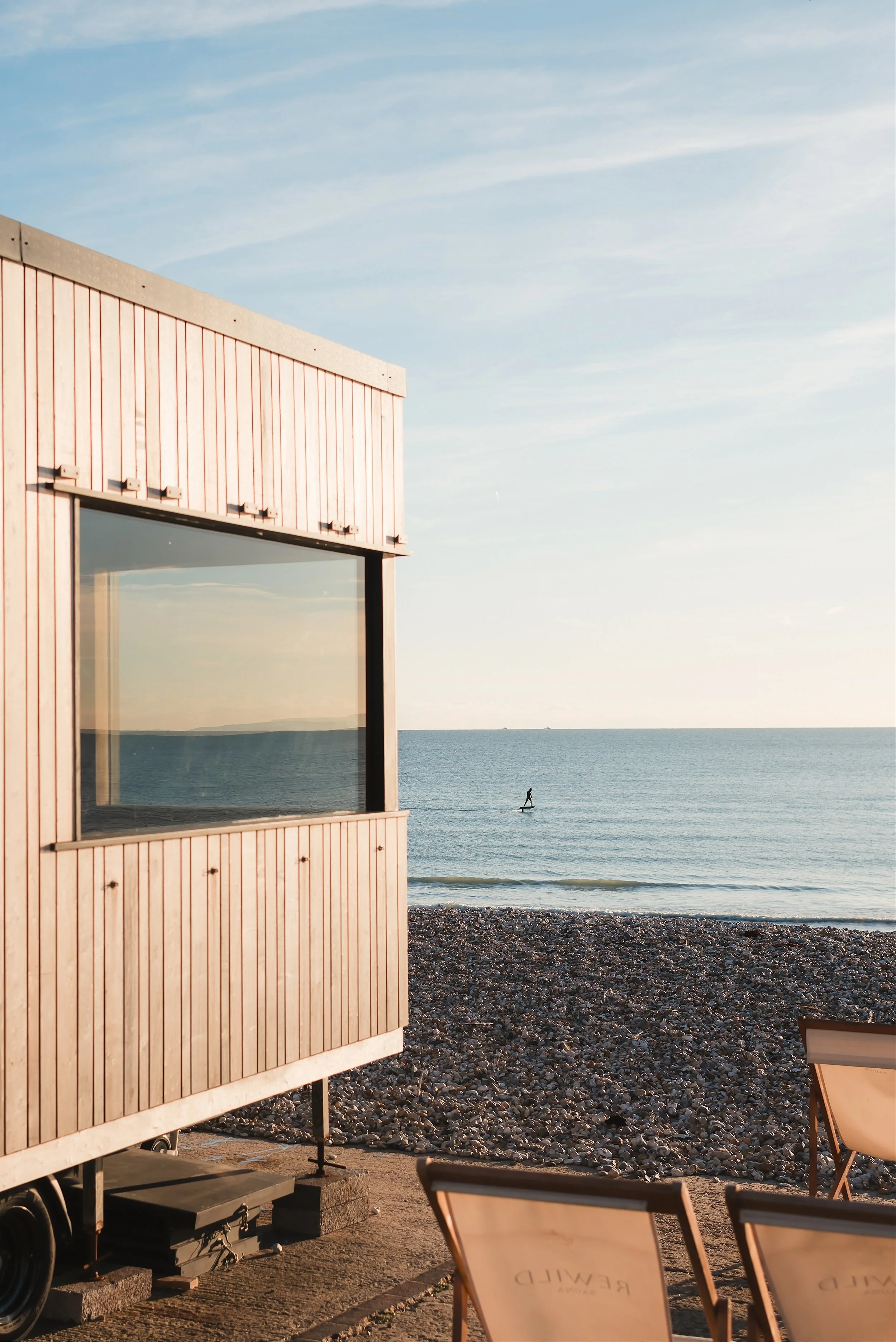 View of a modern building with light wooden exterior and large window overlooking a beach and ocean. A paddleboarder is visible in the water against a cloudy sky.