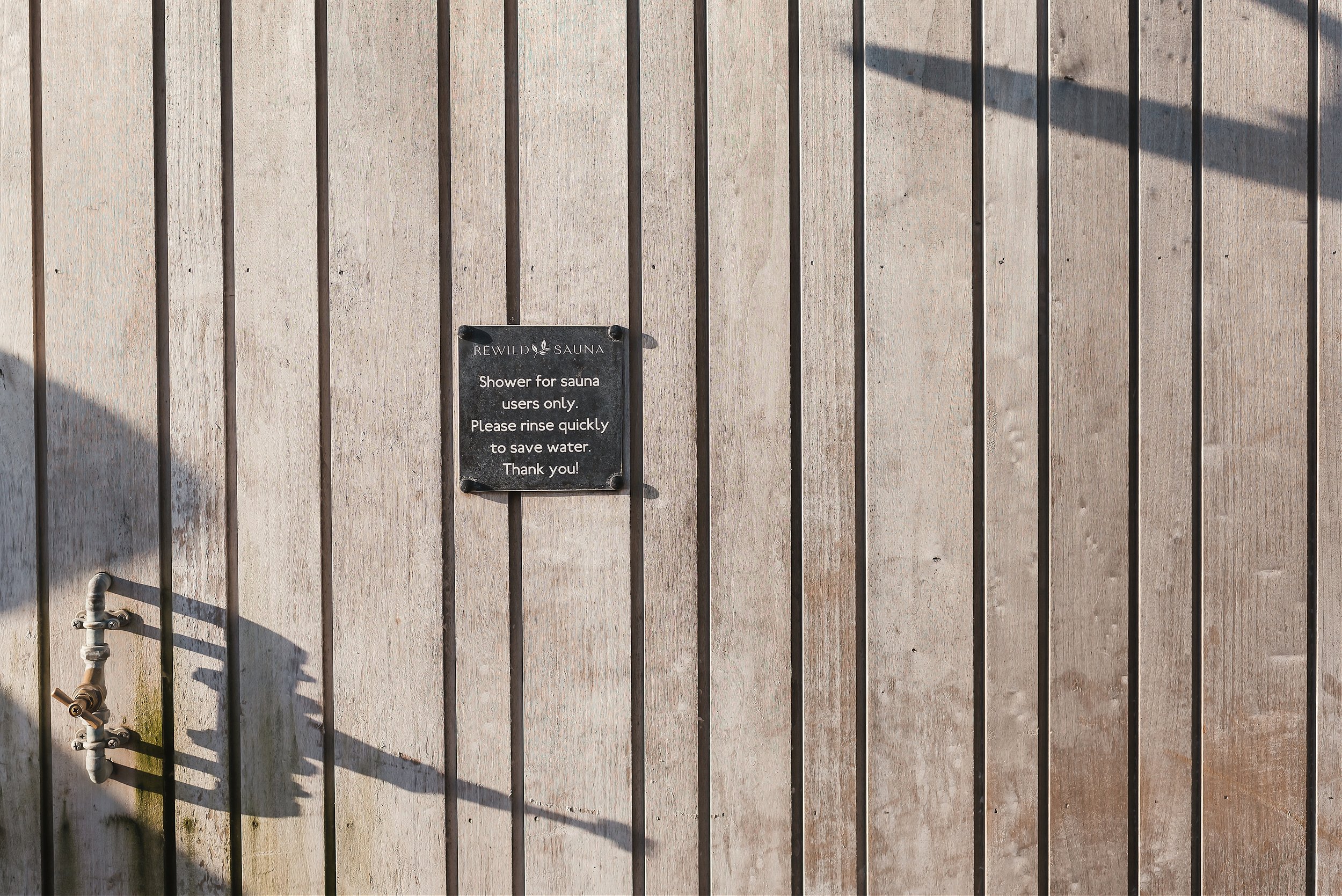 Wooden wall with metal pipes and a black sign that reads 'Rewild Sauna. Shower for sauna users only. Please rinse quickly to save water. Thank you!'