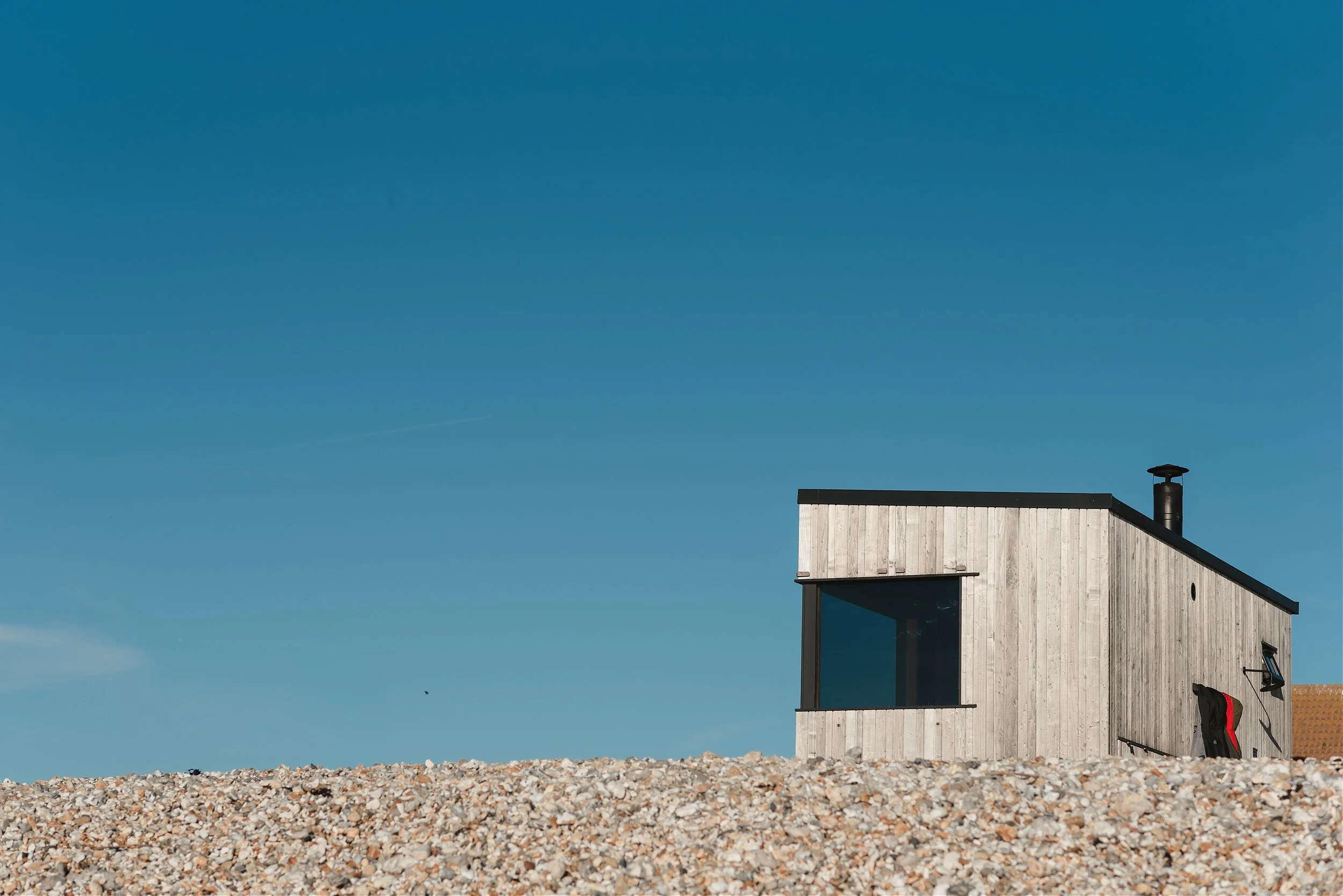 Rewild Sauna wooden cabin with chimney rising above a shingle beach under a clear blue sky in West Sussex