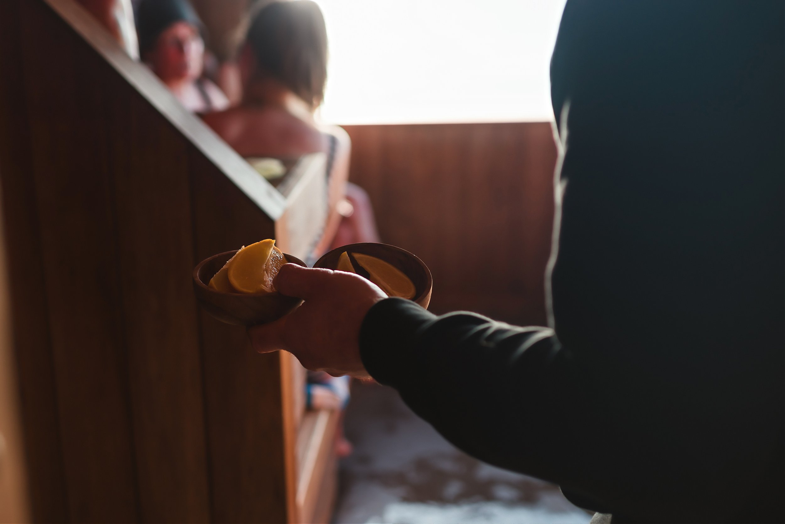 A person in a suit handing over a small bowl with lemon wedges to another person in a black suit, in front of a wooden panel and bright light coming from behind.
