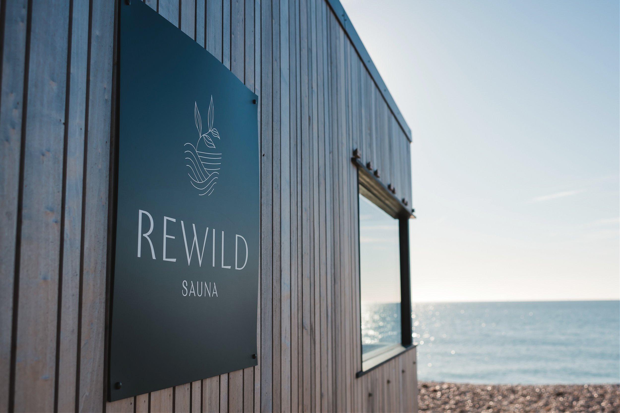 Rewild Sauna branded sign on a timber clad beach sauna cabin with ocean view behind West Sussex