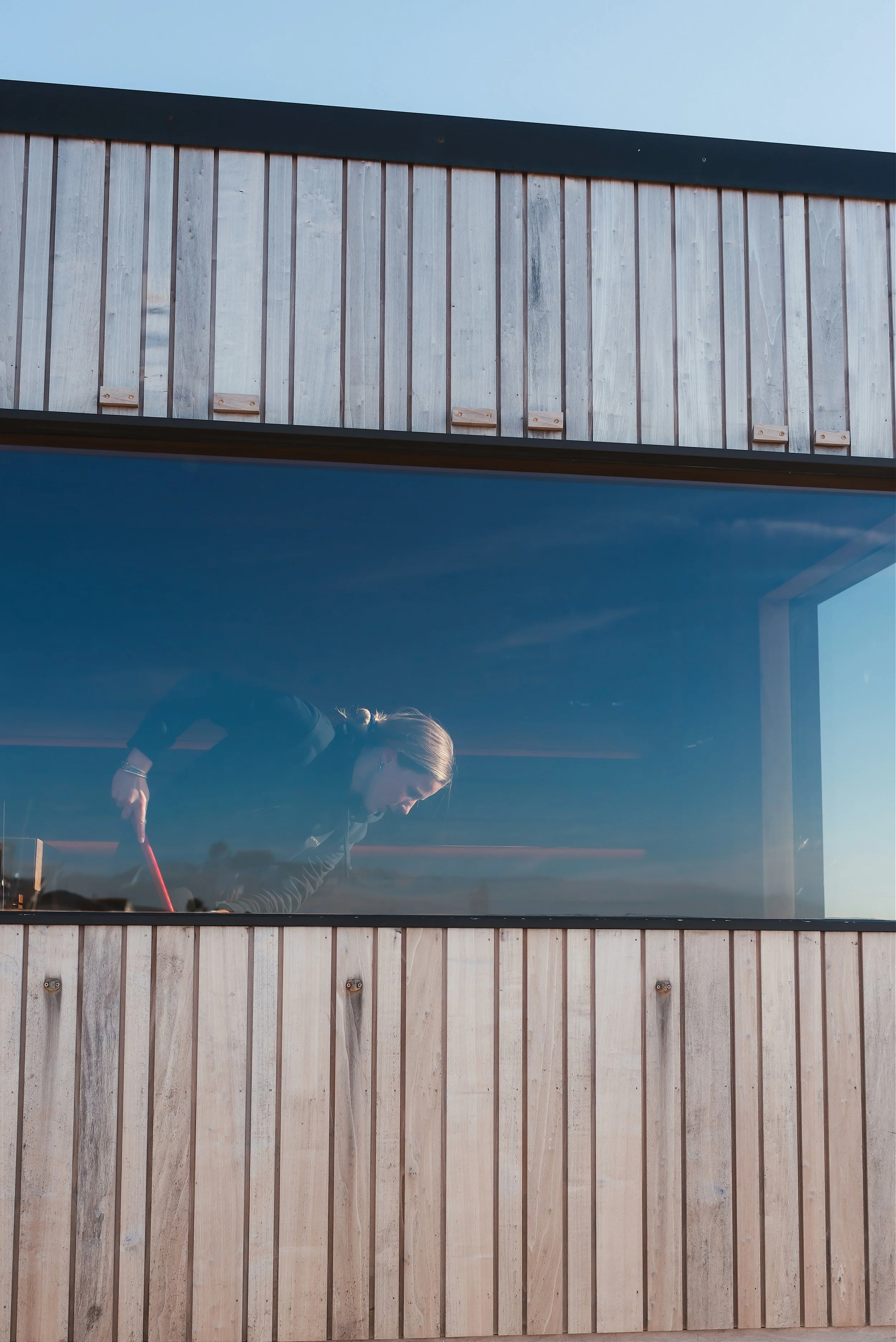 A woman with blonde hair is leaning over a table with a red stick in a room with a large window. The room has a wooden wall and a clear sky is visible outside.