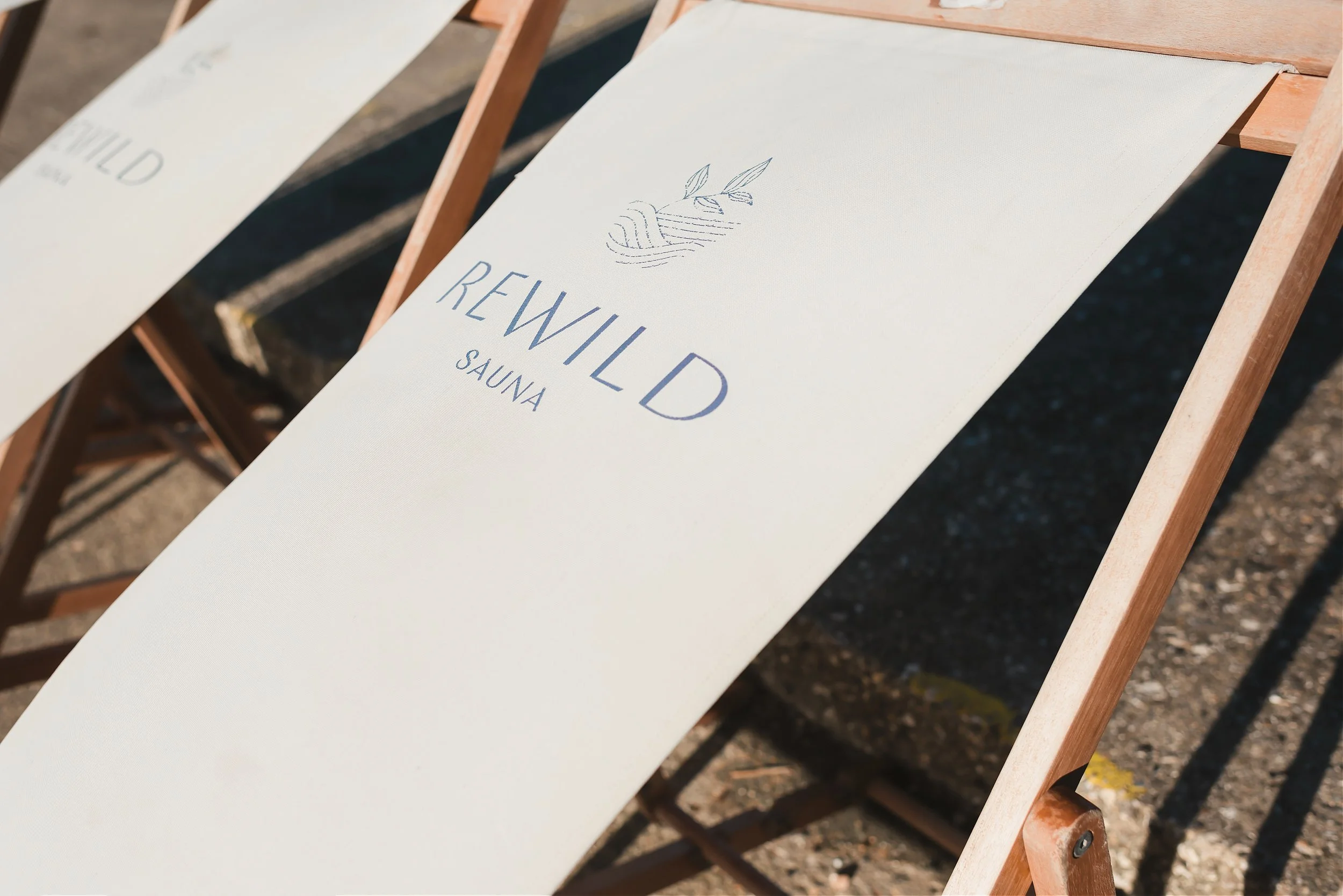Two white lounge chairs with wooden frames, one with a branding that says 'REWILD SAUNA' with an illustration of a beehive and leaves, outdoors on a sunny day.