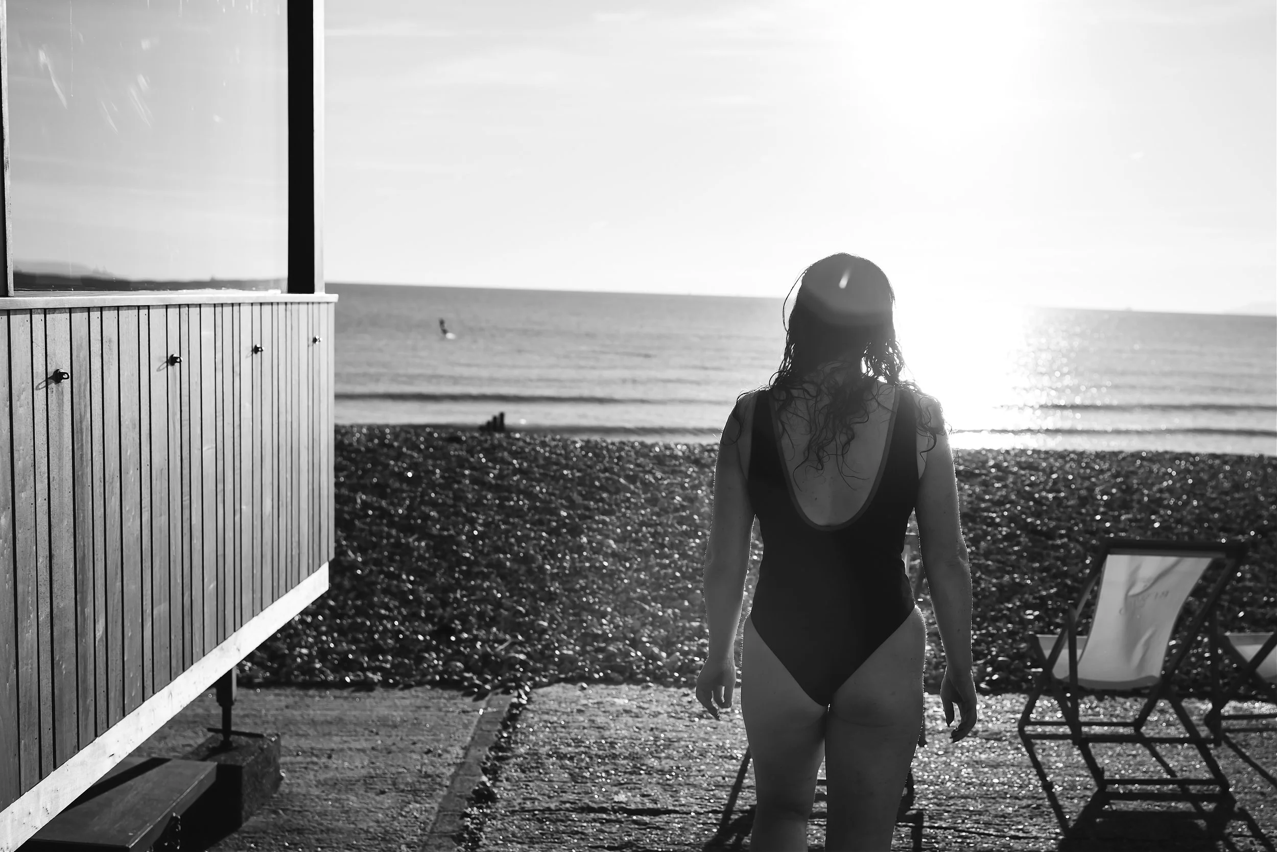 Black and white photo of a woman in a swimsuit walking near a beach as the sun sets over the ocean, with lounge chairs and a wooden building in the foreground.