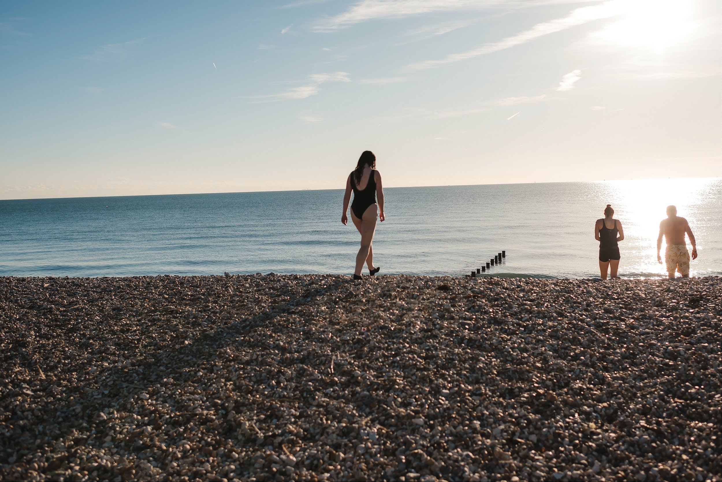 Three people in swimwear walking toward the sea across a sunny Sussex shingle beach for cold water wild swimming