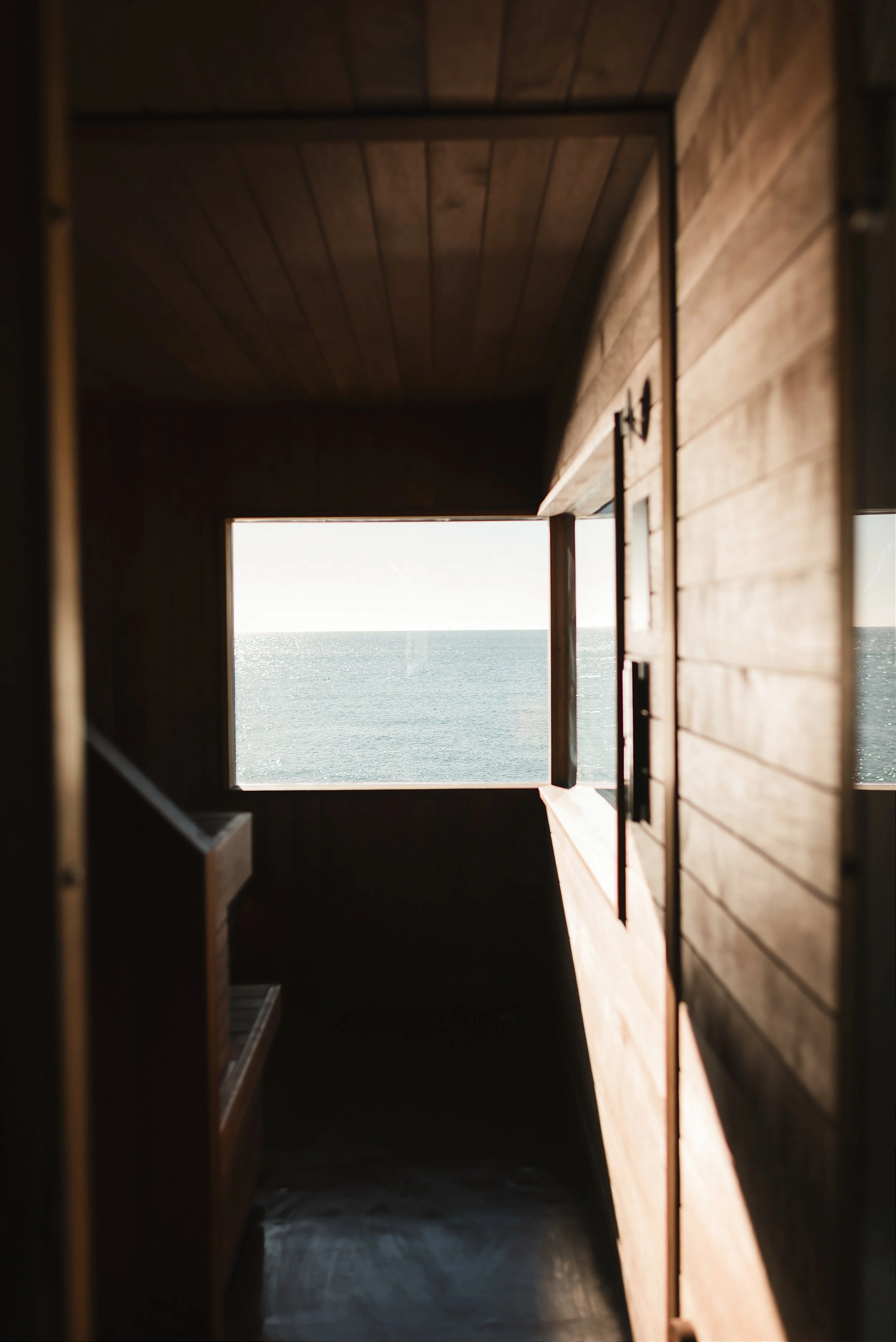 Dark timber interior of a beach sauna with open window framing a glittering sea view West Sussex